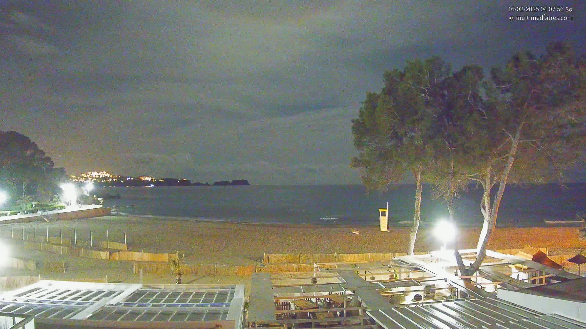 A nighttime view of a beach with calm water under a cloudy sky, shows a sandy shore, a lifeguard stand, and the structure of a beachfront restaurant or bar partially illuminated under the light of several lamps.