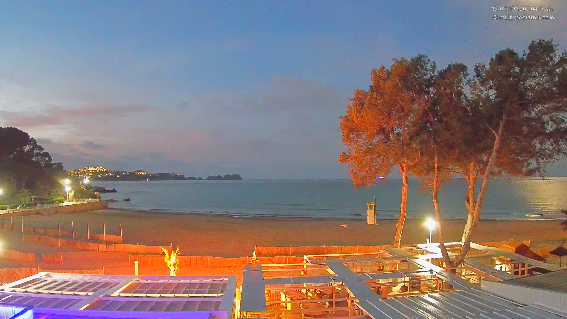 A sandy beach at twilight, with calm ocean water, a beachside structure, and several illuminated trees in the foreground.