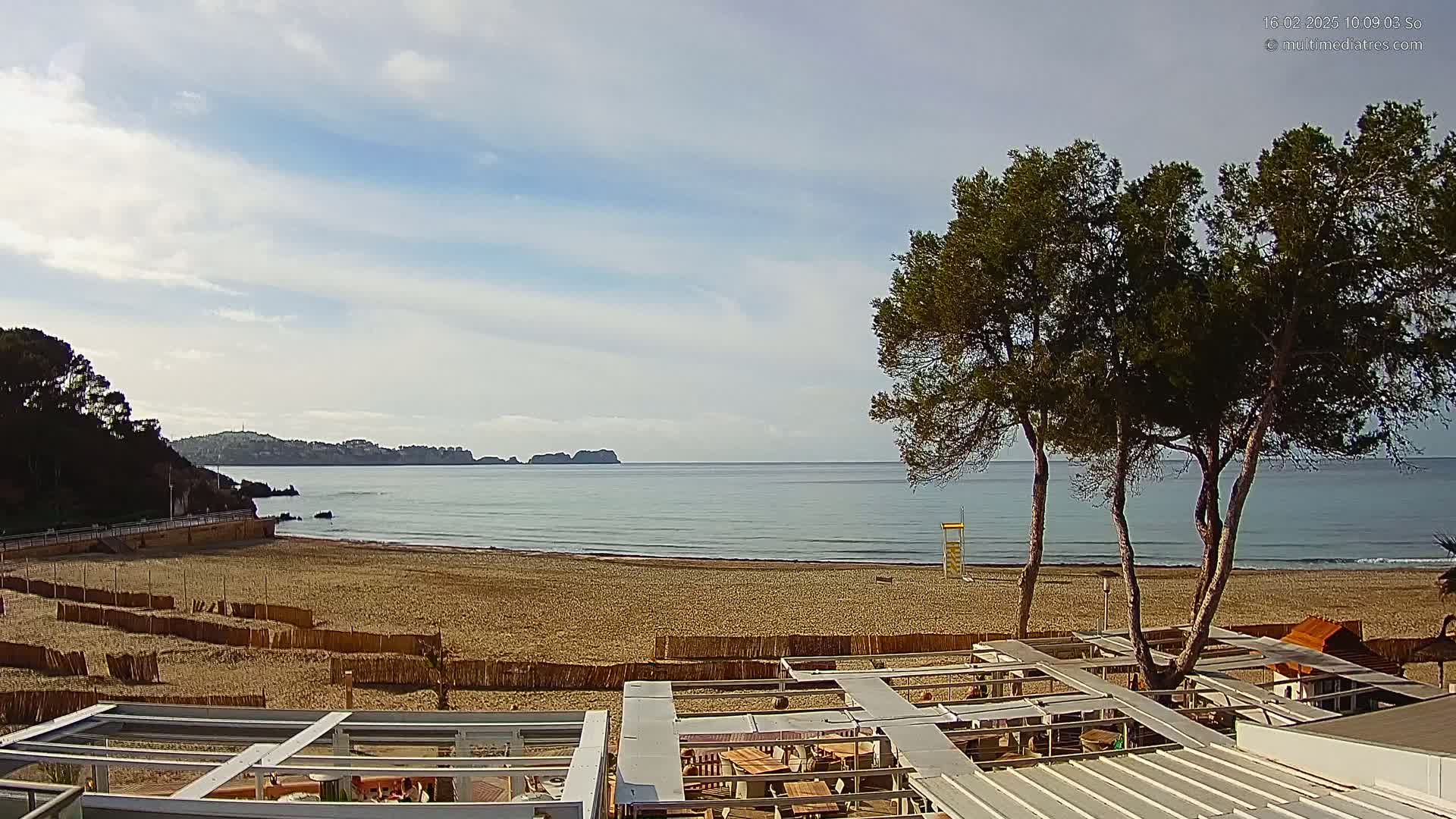 A sandy beach with calm ocean waves stretches to a distant coastline under a partly cloudy sky, with several pine trees standing near a beachfront structure.