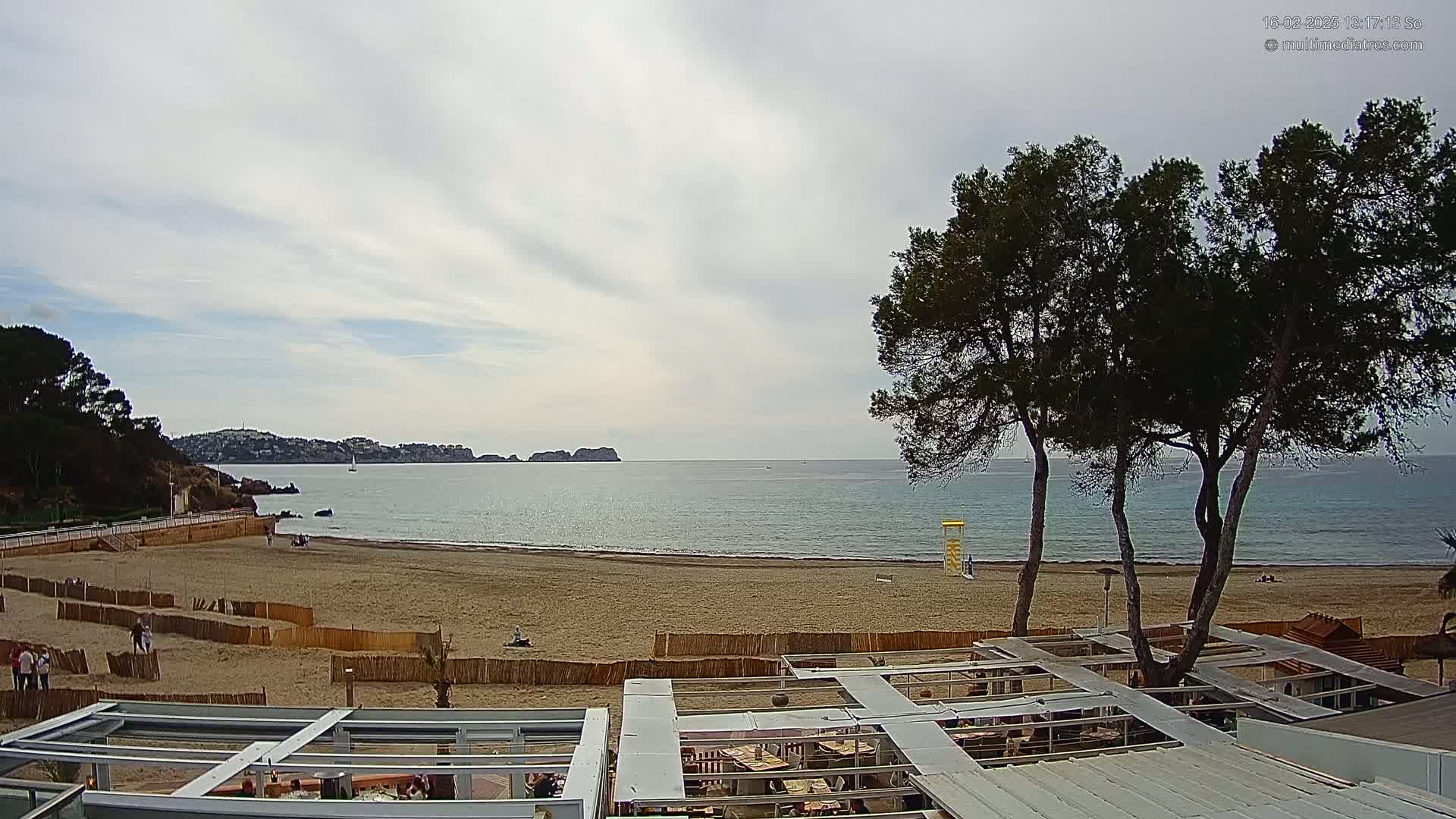 A sandy beach with calm ocean water, a few people, and several pine trees is seen from above a beachside restaurant under a mostly cloudy sky.