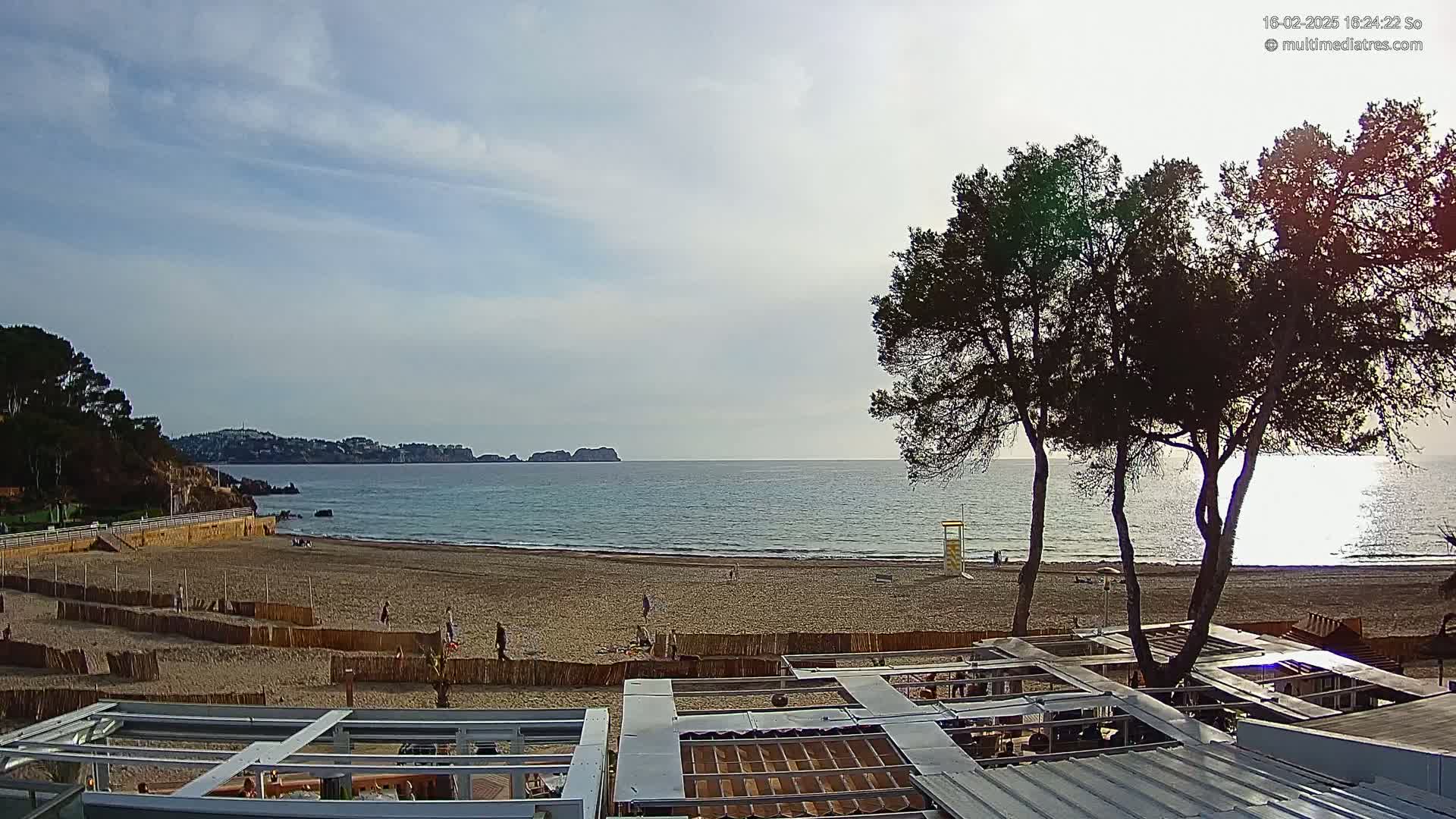 A partly cloudy day at a sandy beach with calm ocean water, overlooking a coastline and distant hills, partially obscured by trees in the foreground.