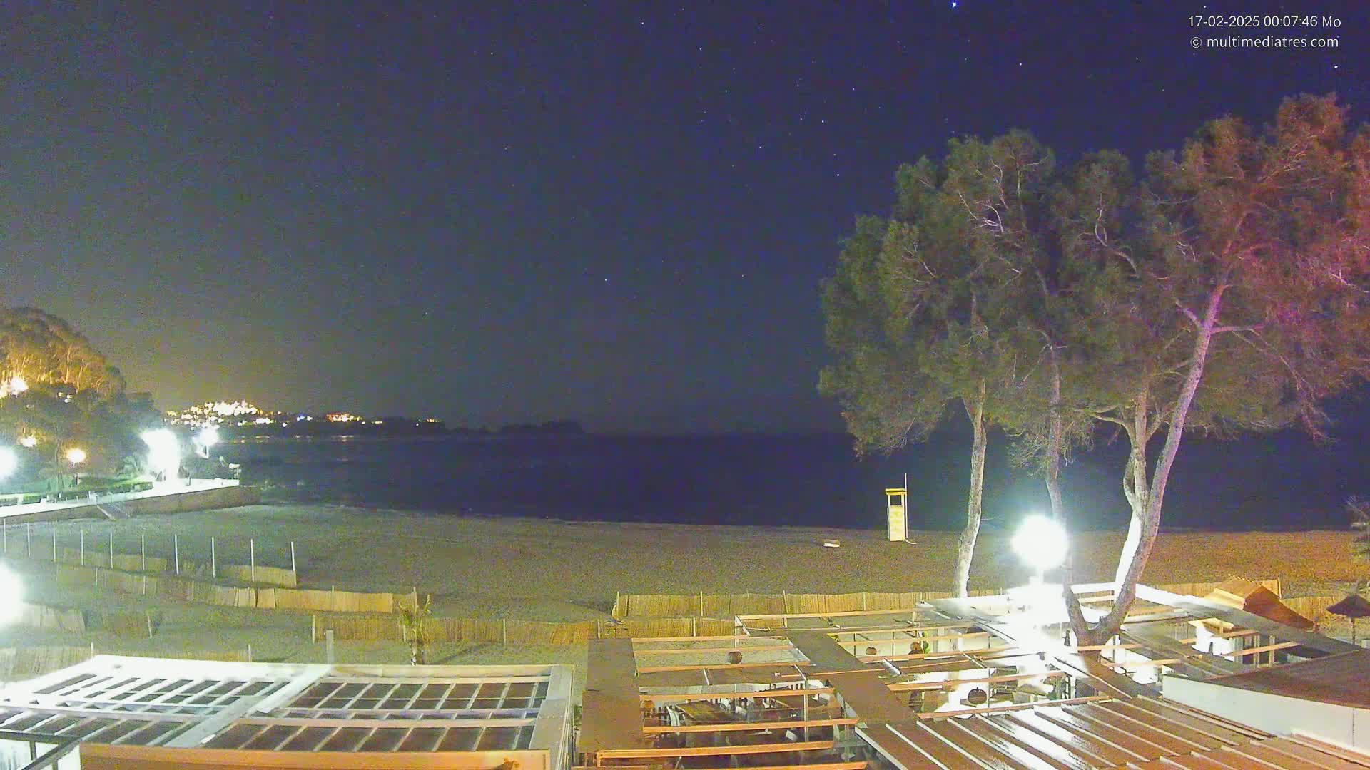 A nighttime beach scene under a clear, starry sky shows a sandy beach, calm water, several tall trees near a building with an open-air patio, and lights illuminating the beachfront area.