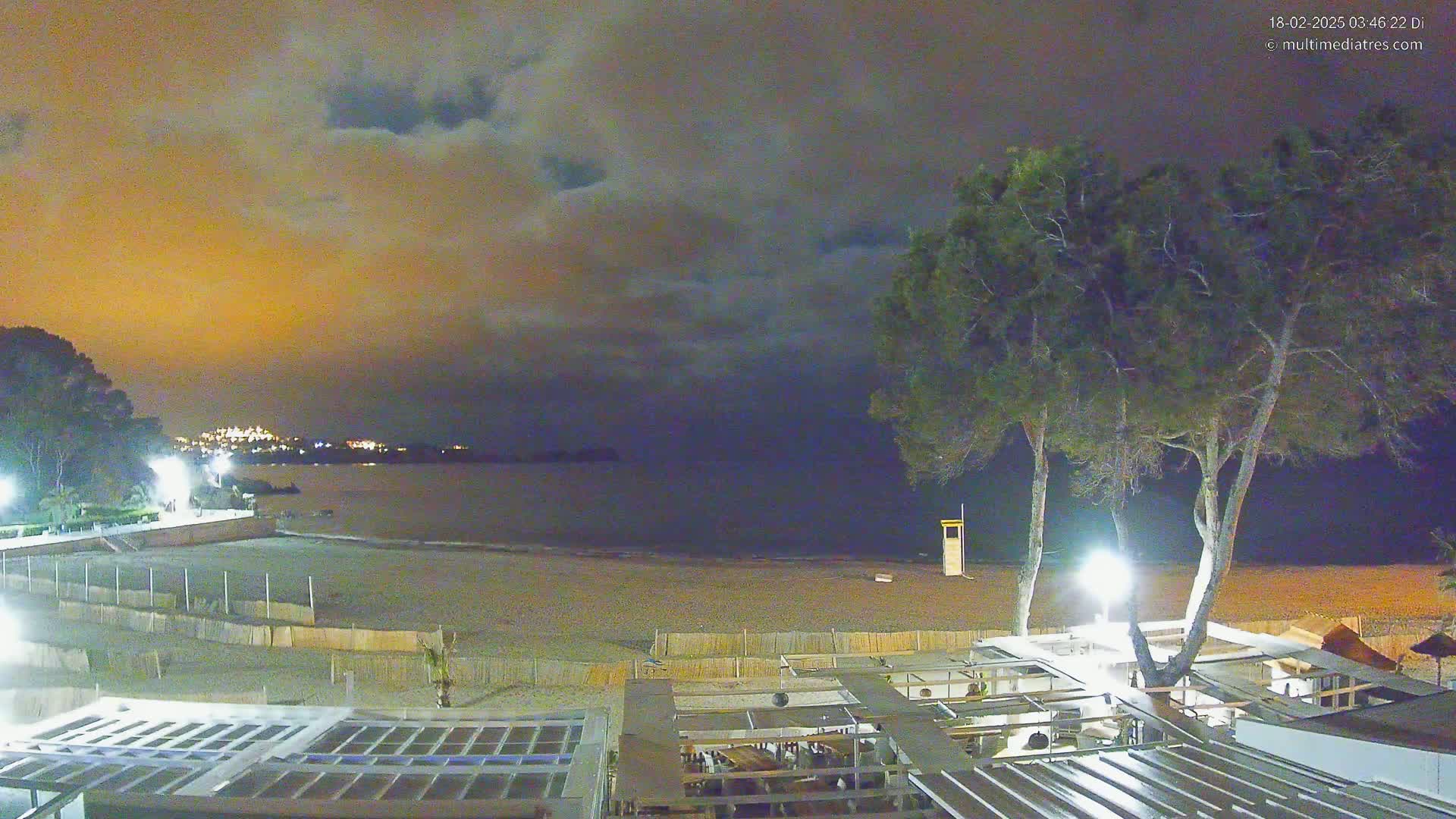 A nighttime beach scene shows a sandy shore, calm water, and a restaurant structure under a cloudy, orange-tinged sky.