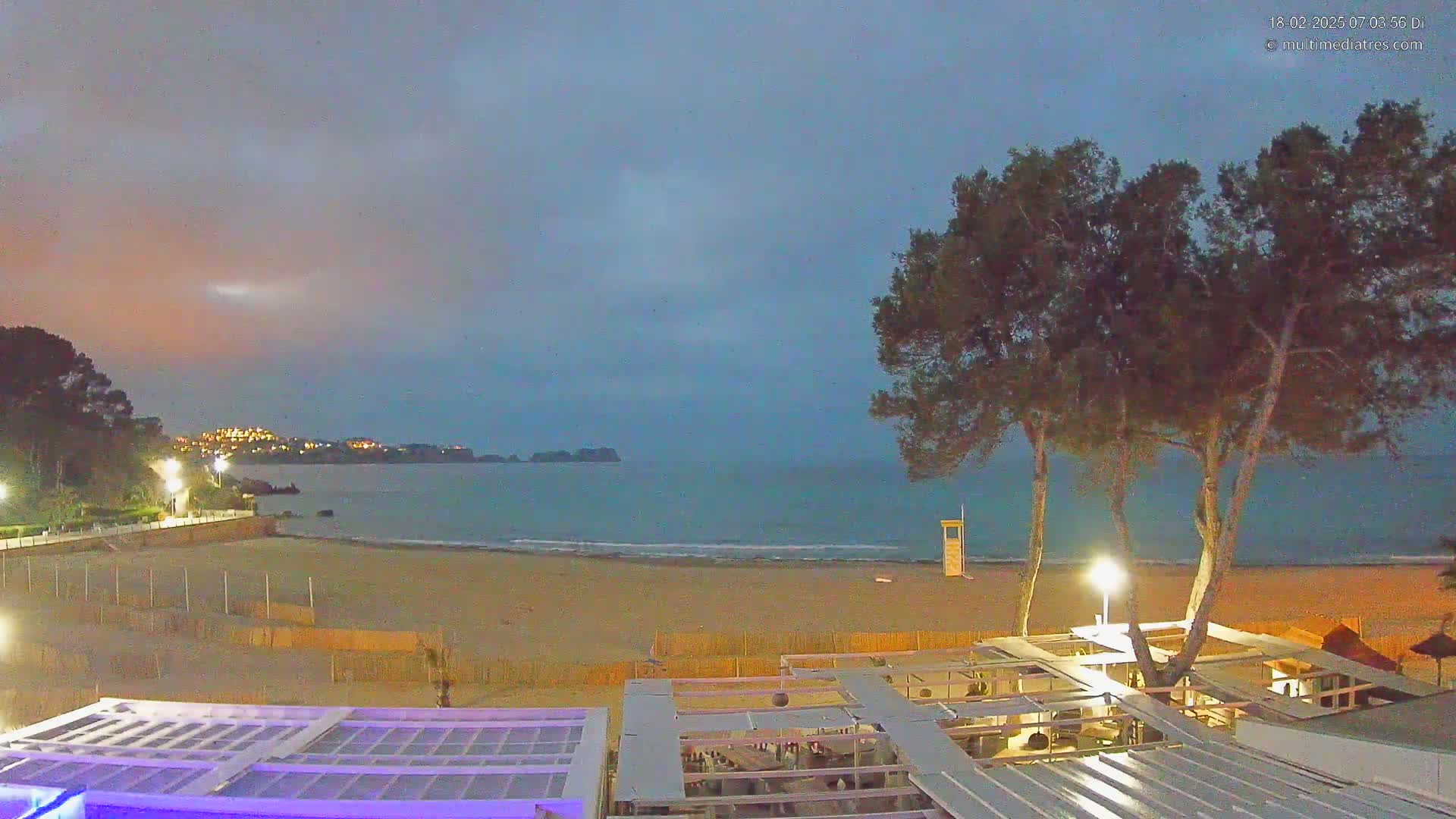 A sandy beach at dusk or dawn is viewed from above, showing a calm sea, a lifeguard stand, a beachfront restaurant with outdoor seating, and tall trees in the foreground under a mostly clear sky.