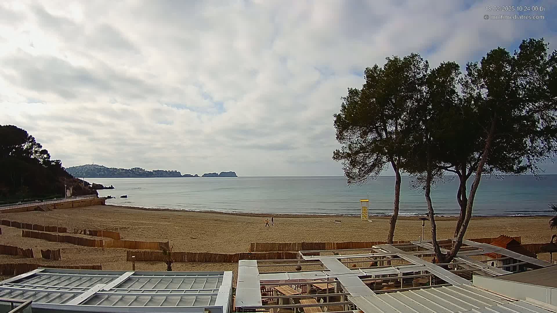 A mostly cloudy day reveals a sandy beach with calm ocean waters, a distant coastline, and several pine trees near a beachfront structure.