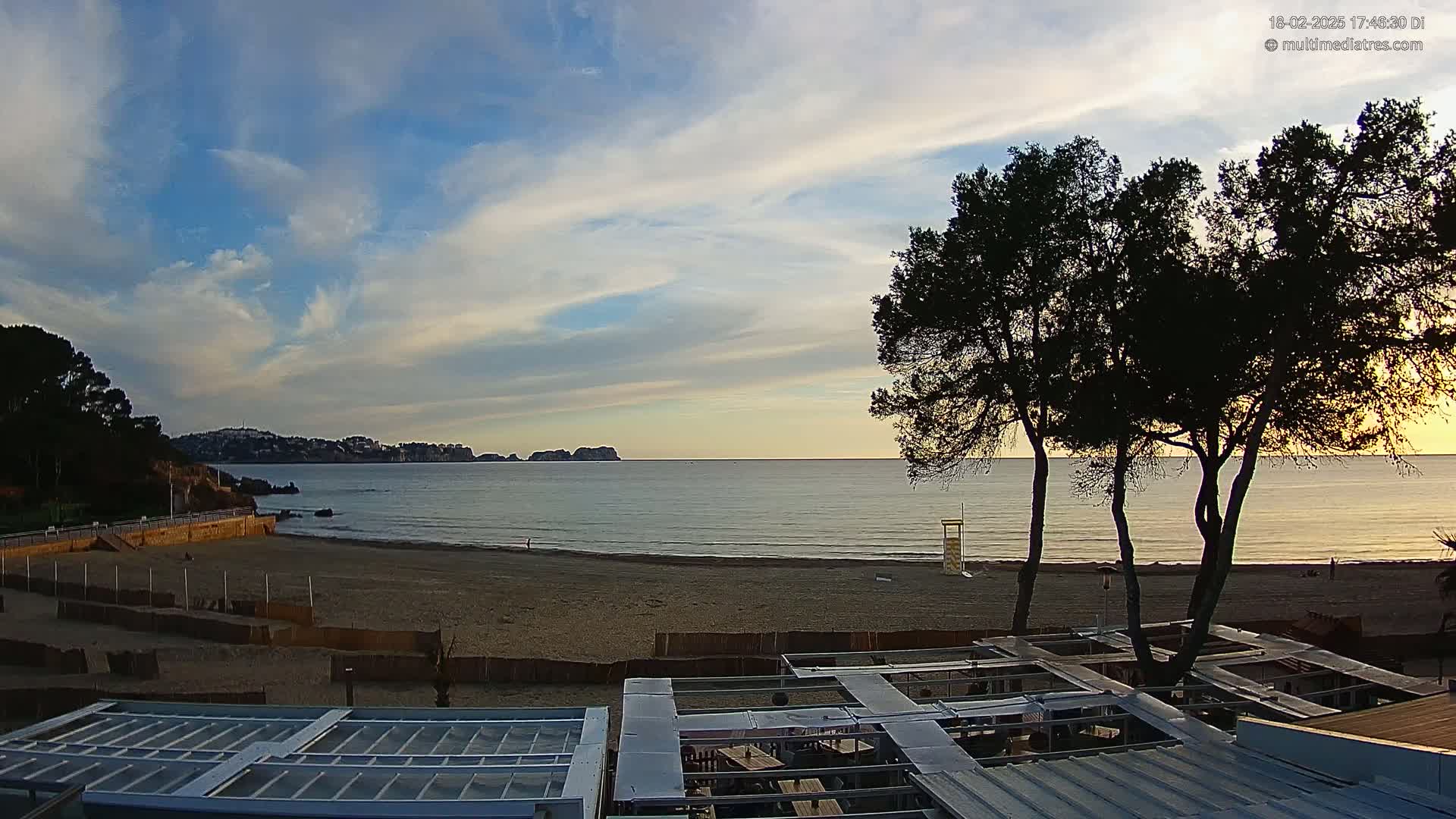 A sandy beach with calm ocean water under a partly cloudy sky is viewed from above a beachside structure at sunset.