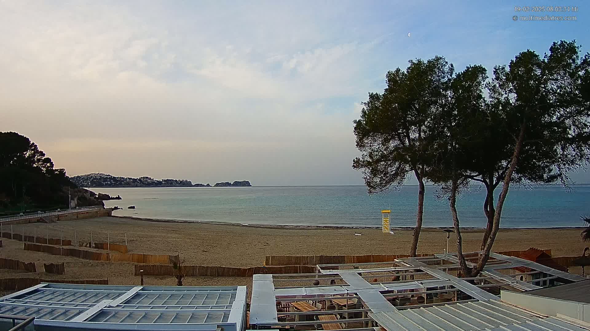 A sandy beach with calm, blue-green water stretches out under a mostly cloudy sky, overlooked by a seaside restaurant and several tall pine trees.