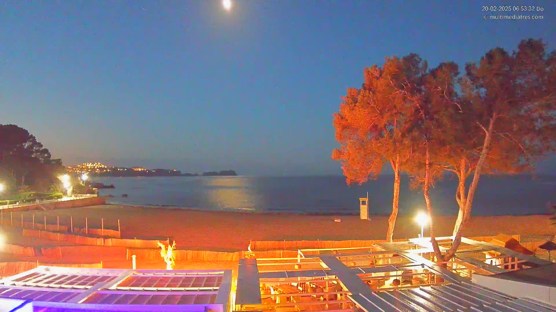 A moonlit beach at night, with a calm sea, illuminated buildings in the distance, and a beachfront structure with outdoor seating.