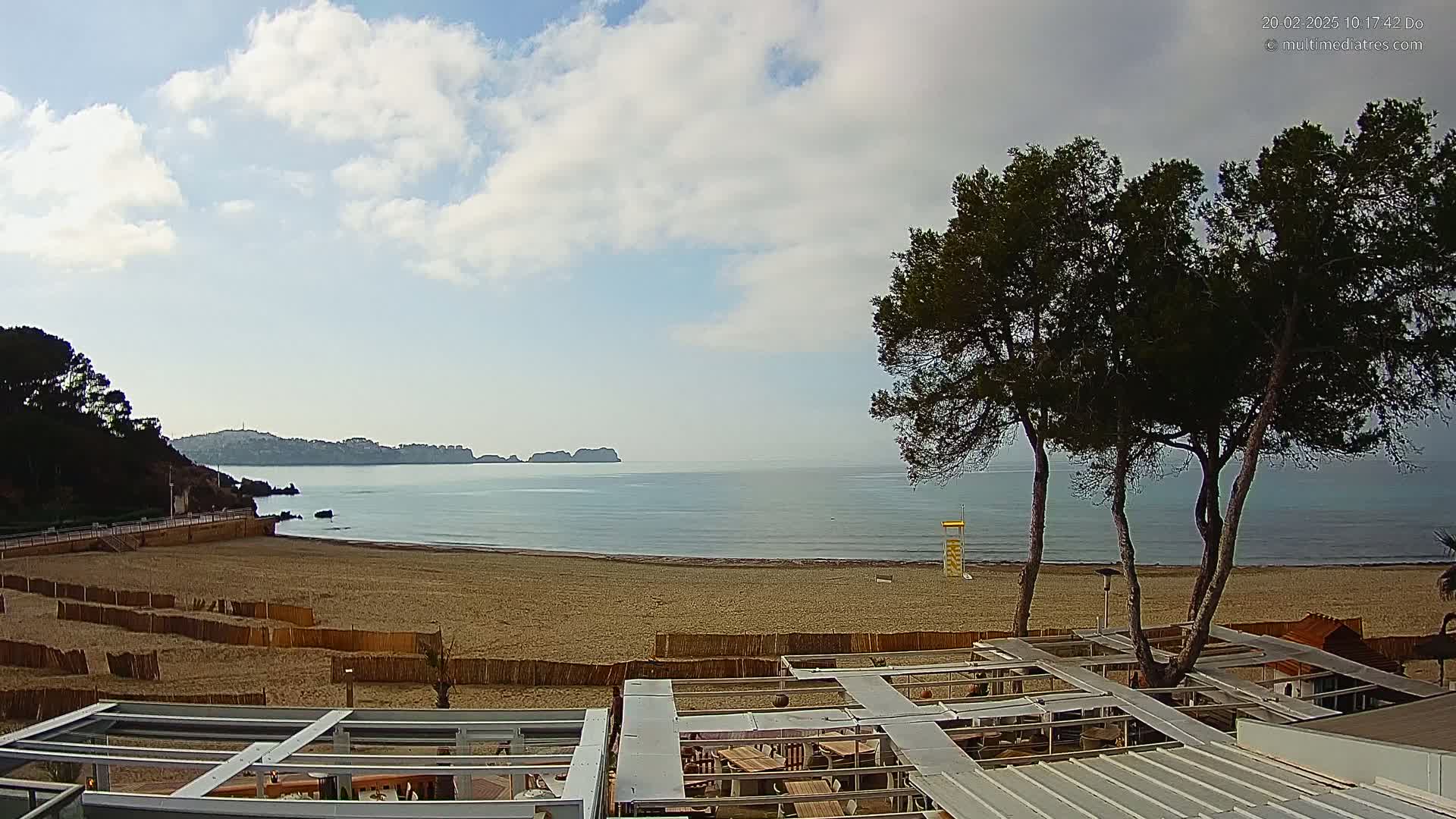 A sandy beach with calm ocean water, partially cloudy skies, and several pine trees in the foreground, all viewed from atop a structure with a metal roof.