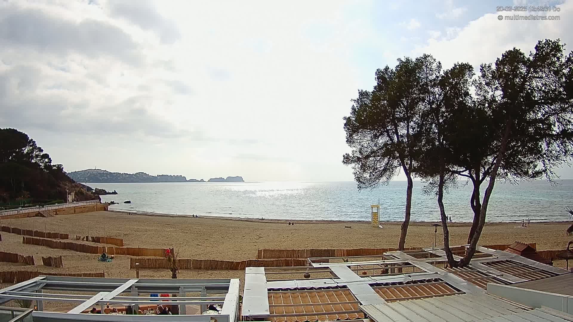 A sandy beach with calm, somewhat cloudy, blue water stretches to a distant coastline under a partly cloudy sky, overlooked by several pine trees and beachside structures.