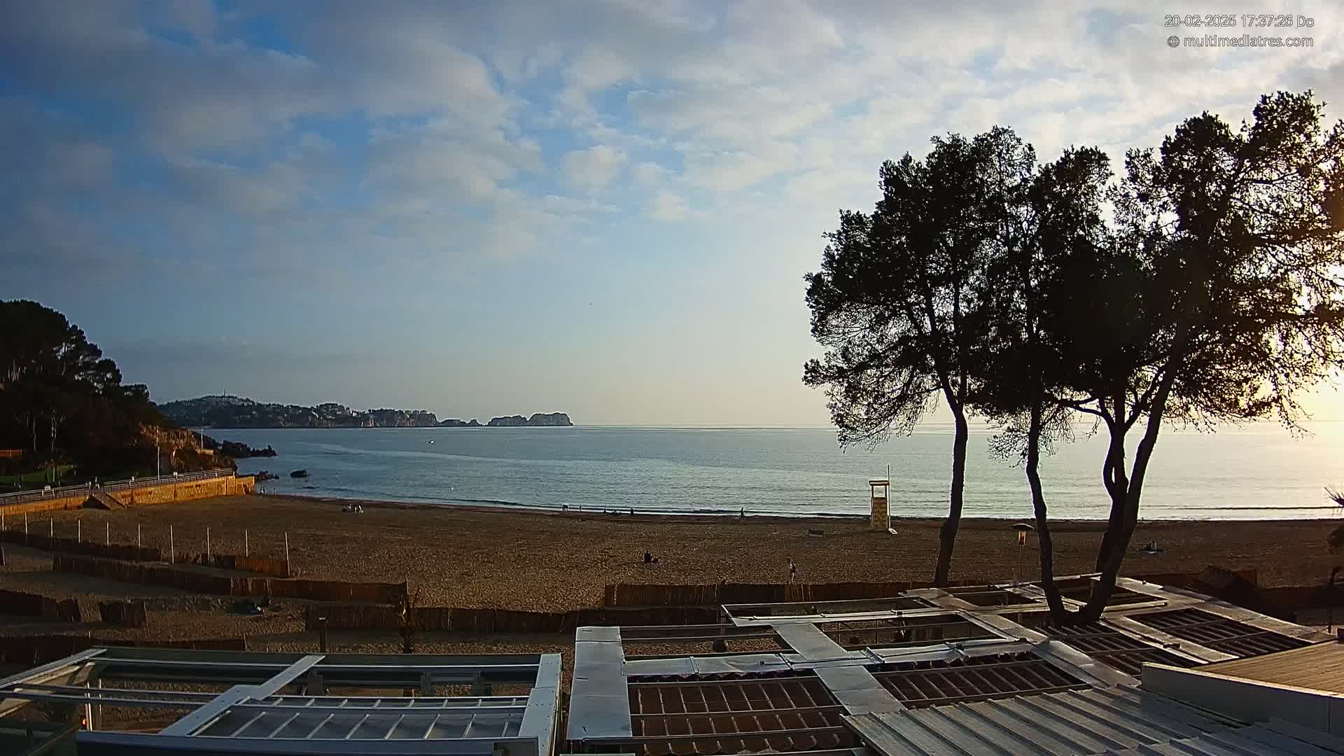 A sandy beach stretches towards a calm sea under a partly cloudy sky, with dark trees silhouetted in the foreground.