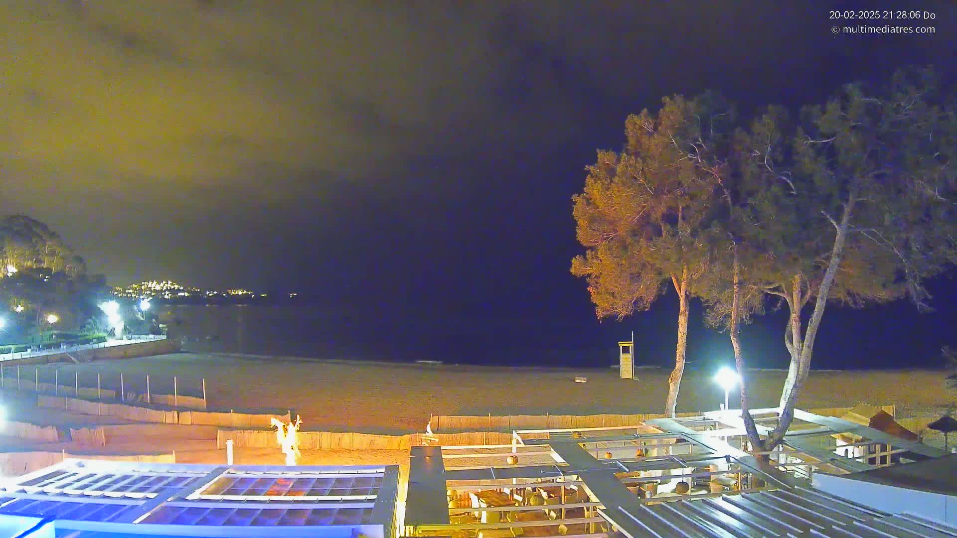 A nighttime view of a beach at night with a few trees and an outdoor restaurant, under a partly cloudy sky.