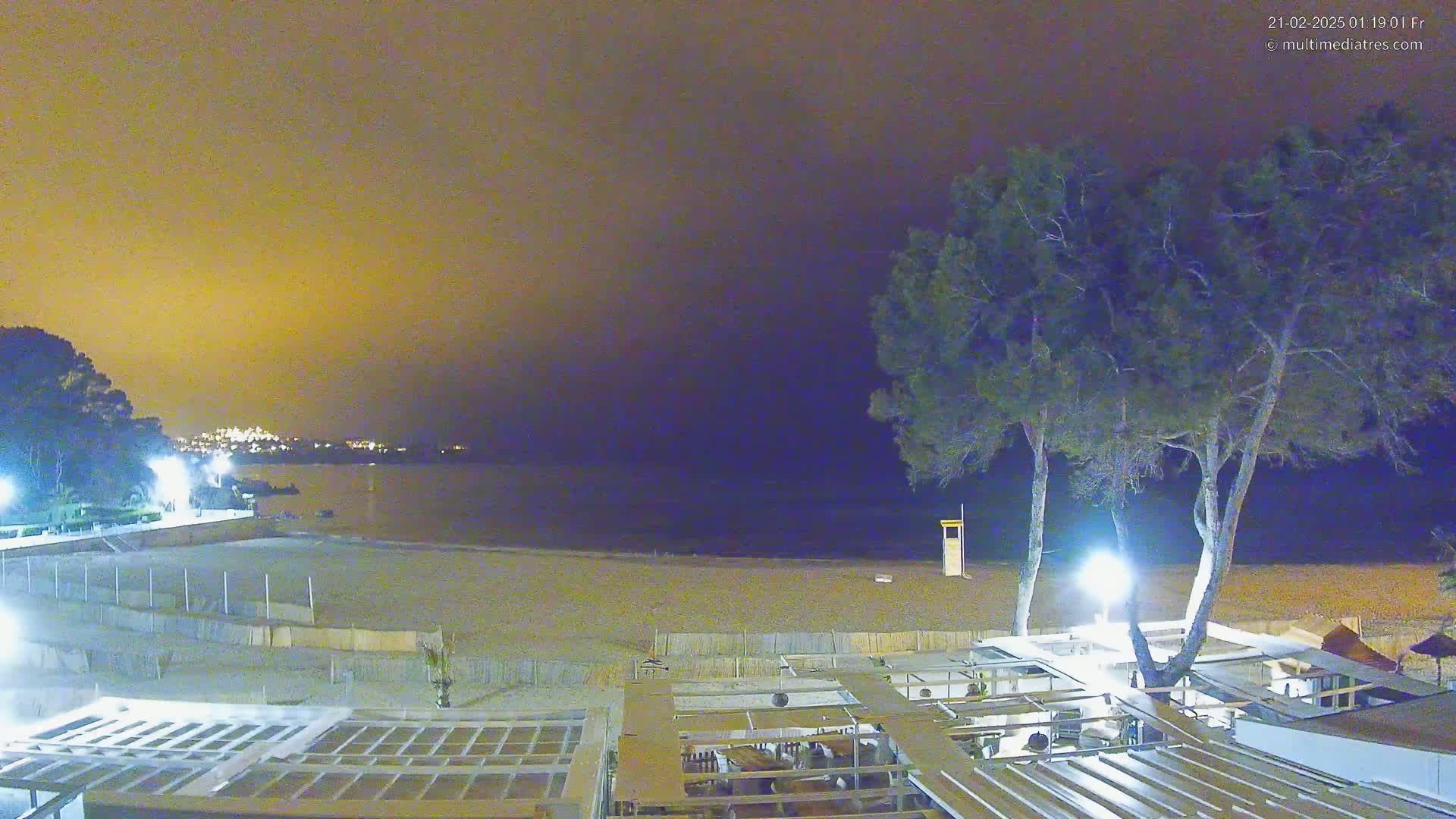 A nighttime view of a beach with a sandy shore, calm water, and a restaurant under a hazy, yellow-tinged sky.
