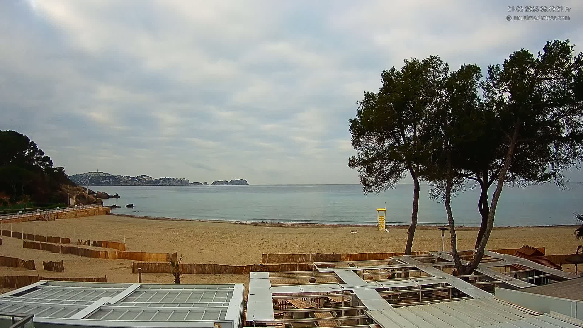 A sandy beach with calm water and a cloudy sky is seen from above a partially covered outdoor seating area, with several pine trees in the foreground.