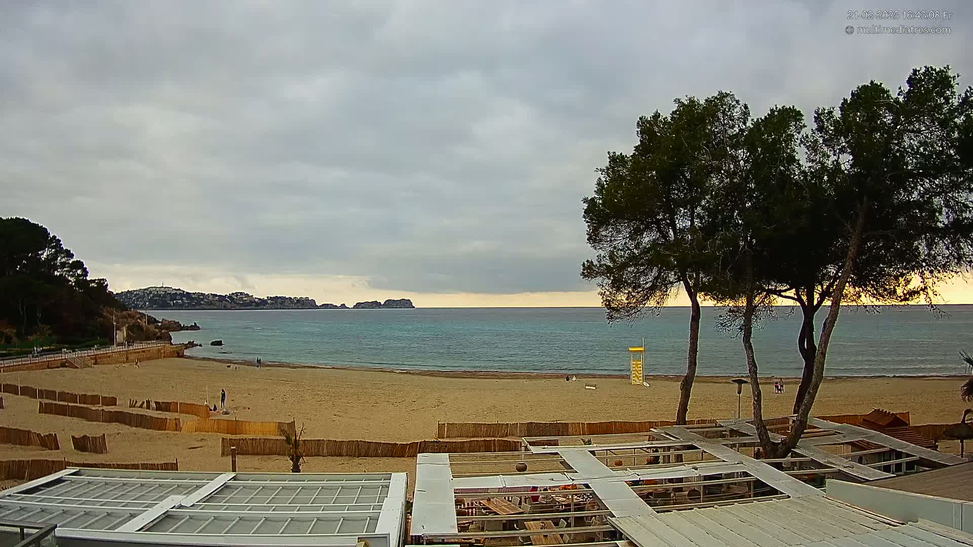 A sandy beach with calm, turquoise water and a few people is visible under a cloudy sky, with trees and beachside structures in the foreground.