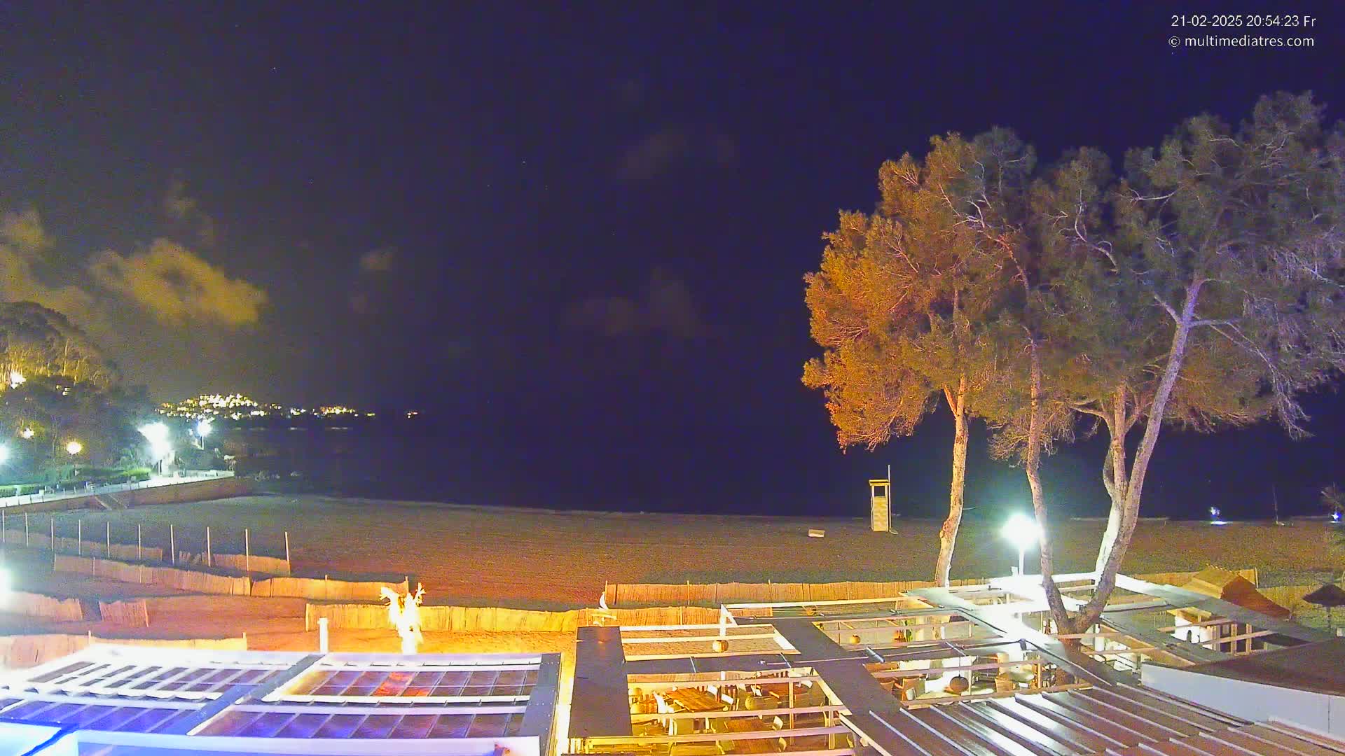 A nighttime view of a beach with a partly cloudy sky, illuminated by artificial lights, showing a sandy beach, a beachfront restaurant, and several trees.