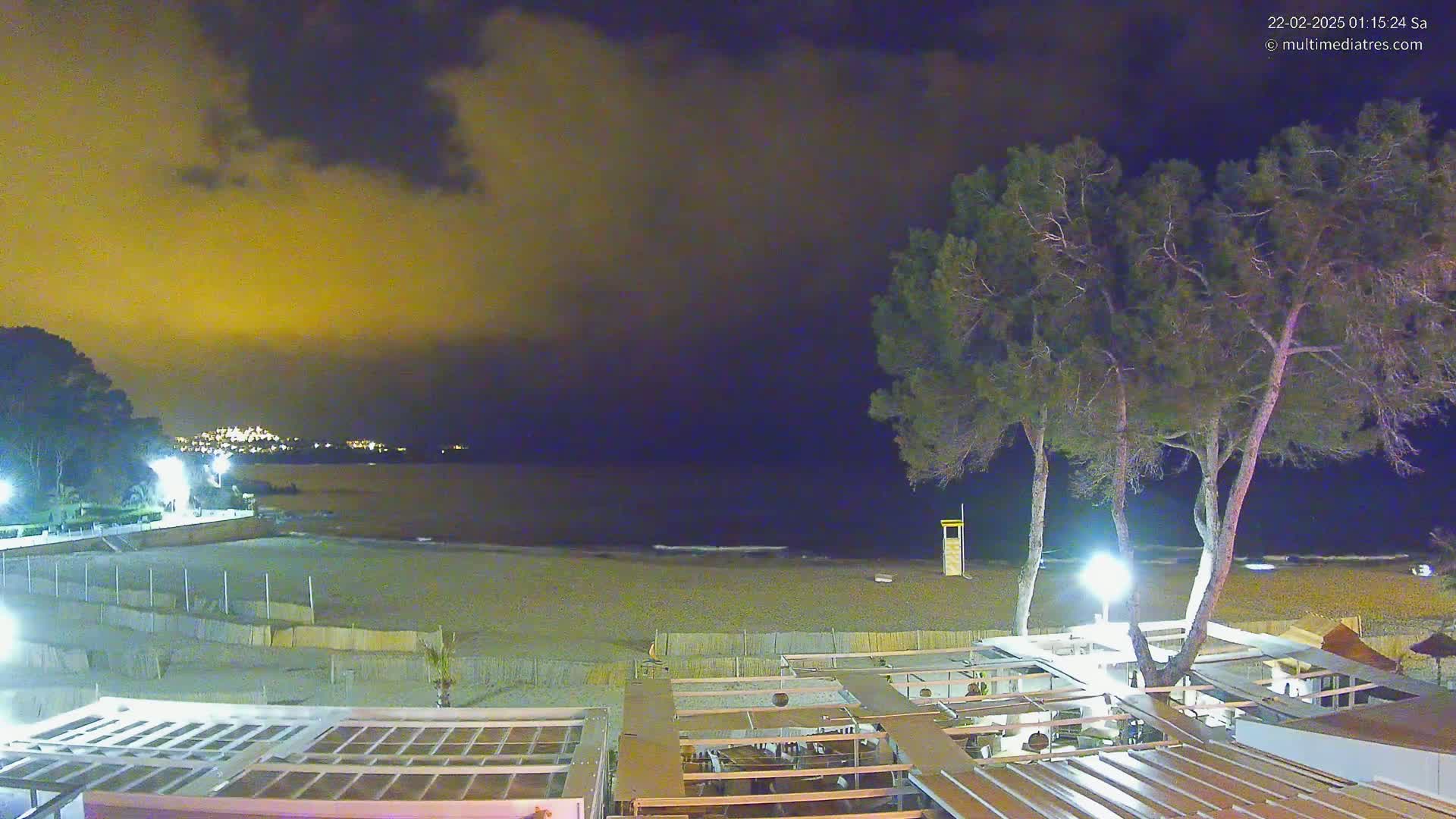 A nighttime view of a beach with a partially cloudy, dark yellow sky, showing a sandy beach, a shoreline with buildings, trees, and lights.