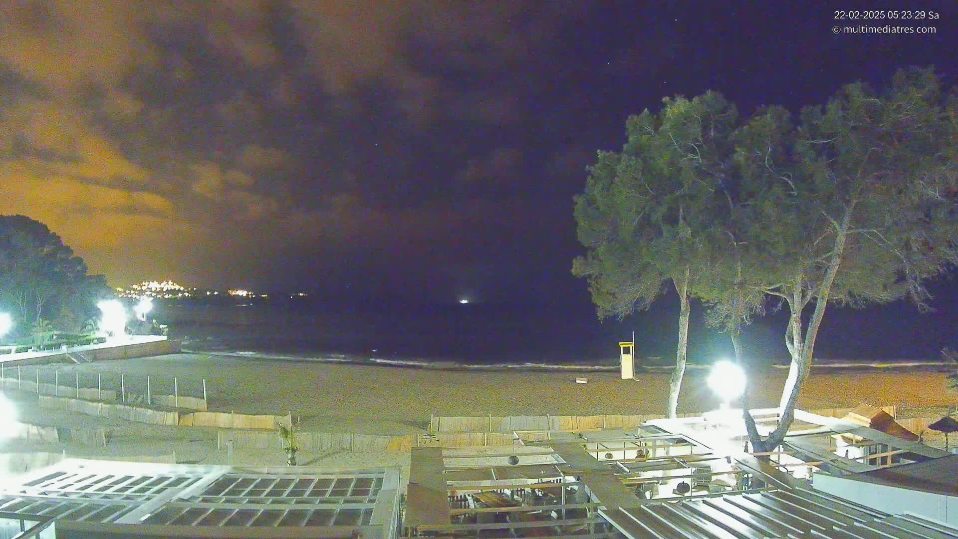 A nighttime beach scene under a cloudy sky shows a sandy beach, calm ocean waves,  trees with lights illuminating a beachfront building, and distant lights on a coastline.