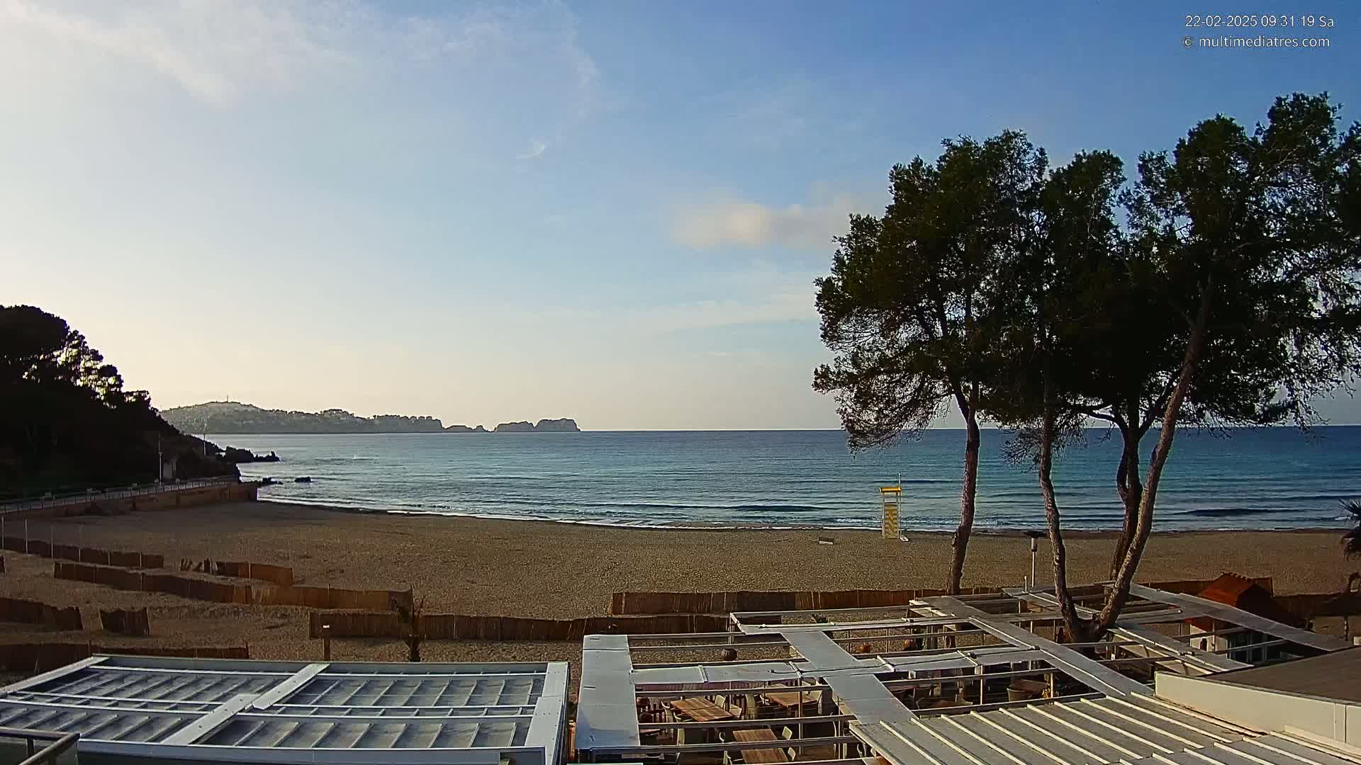 A sandy beach with calm, light-blue water stretches to a distant coastline under a clear, bright sky; several trees stand between the beach and a partially covered outdoor structure.
