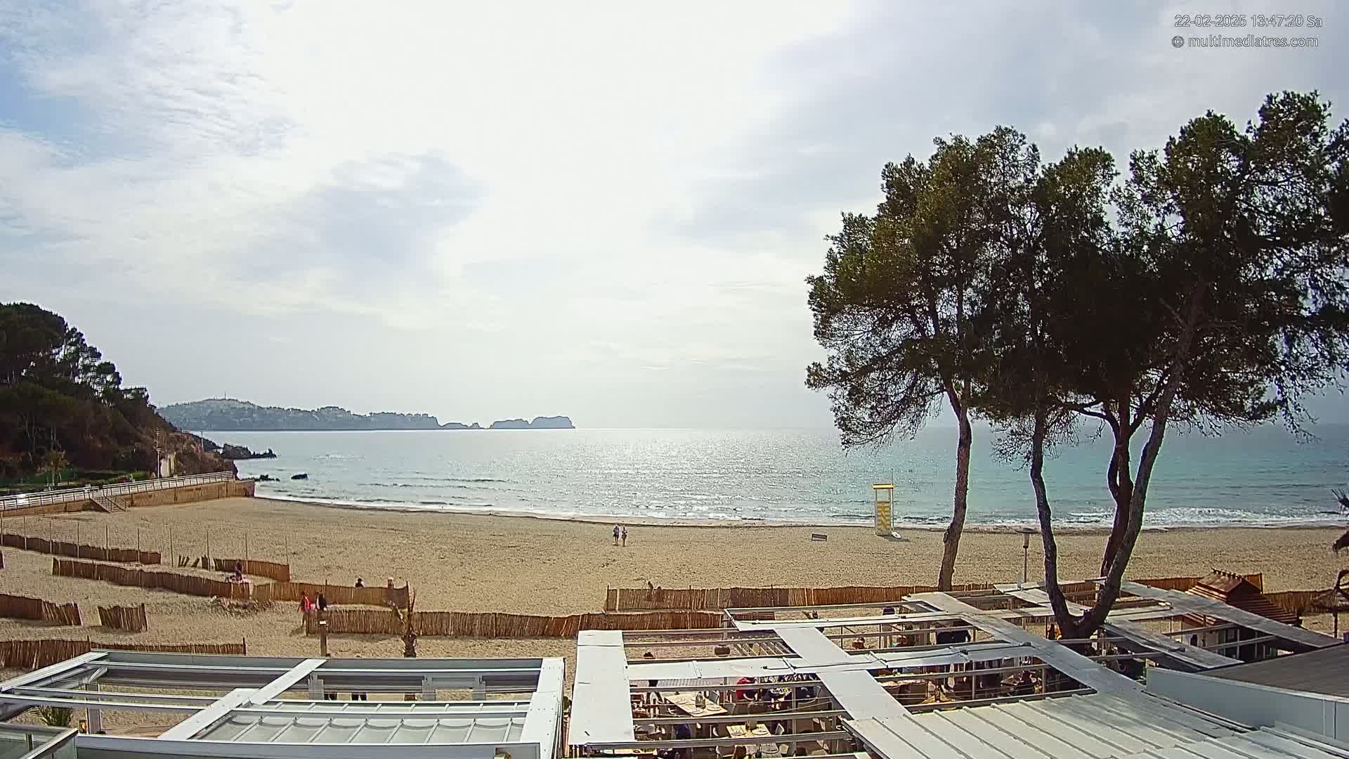 A sandy beach with calm waves under a partly cloudy sky is seen from above a beachfront restaurant.