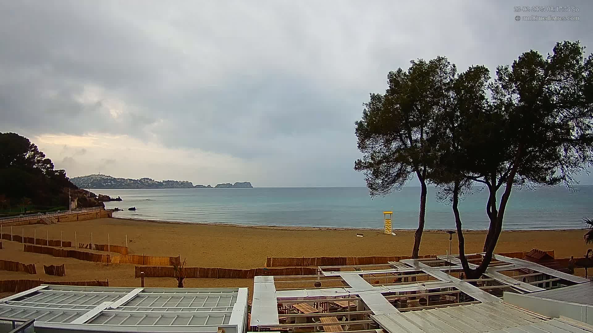 A sandy beach with calm, light blue water under an overcast sky is partially enclosed by wooden fences, with a lifeguard stand visible in the distance and several trees in the foreground.
