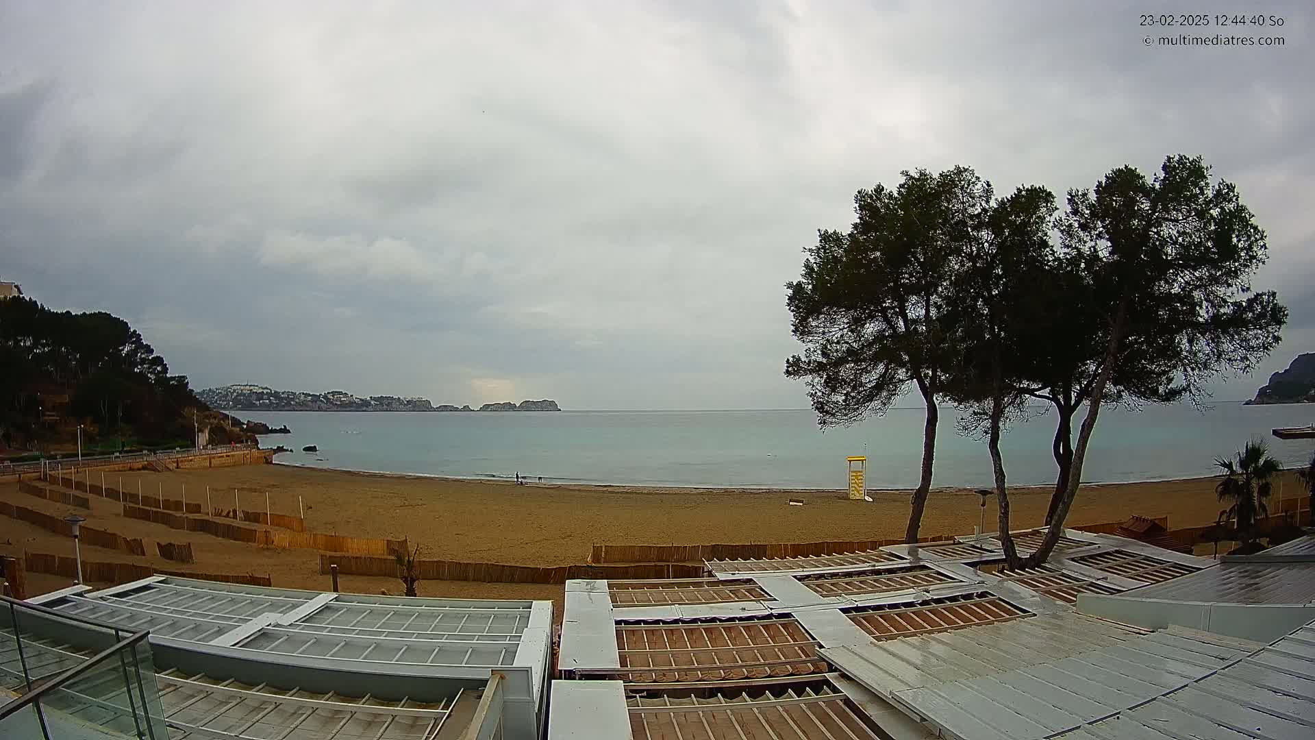 A sandy beach with calm, light-blue water under an overcast sky, overlooked by several pine trees and a building with retractable roofs.