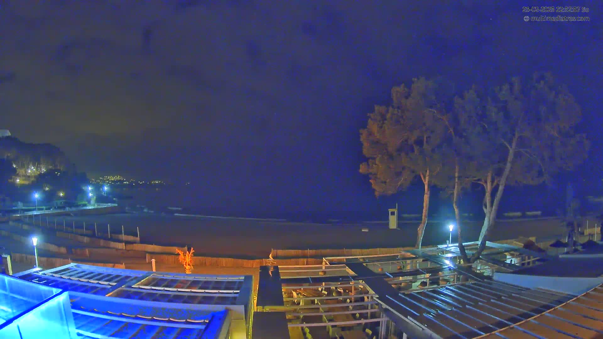 An elevated nighttime view of a beach, with a dark sky, showing a restaurant structure and two trees near the shore.