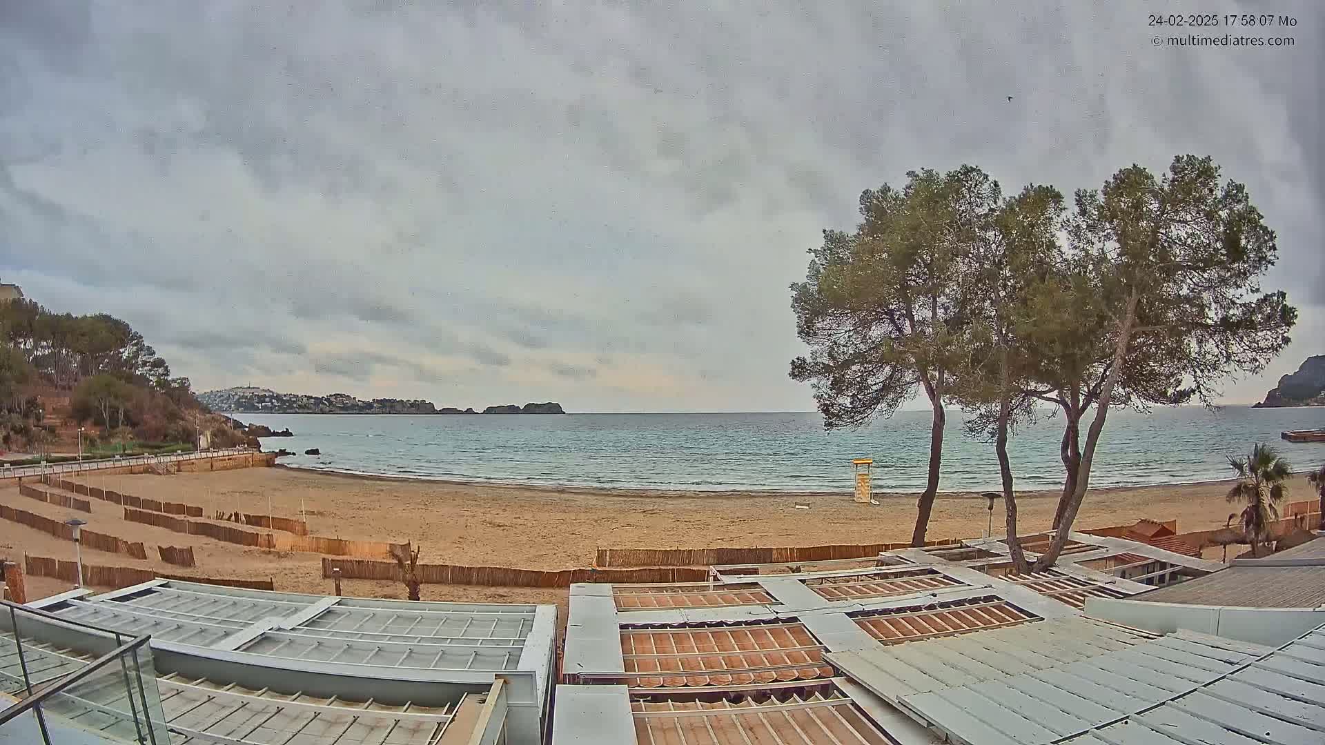 An overcast sky hangs over a sandy beach with calm water, several pine trees, and the roofs of beachside buildings visible in the foreground.