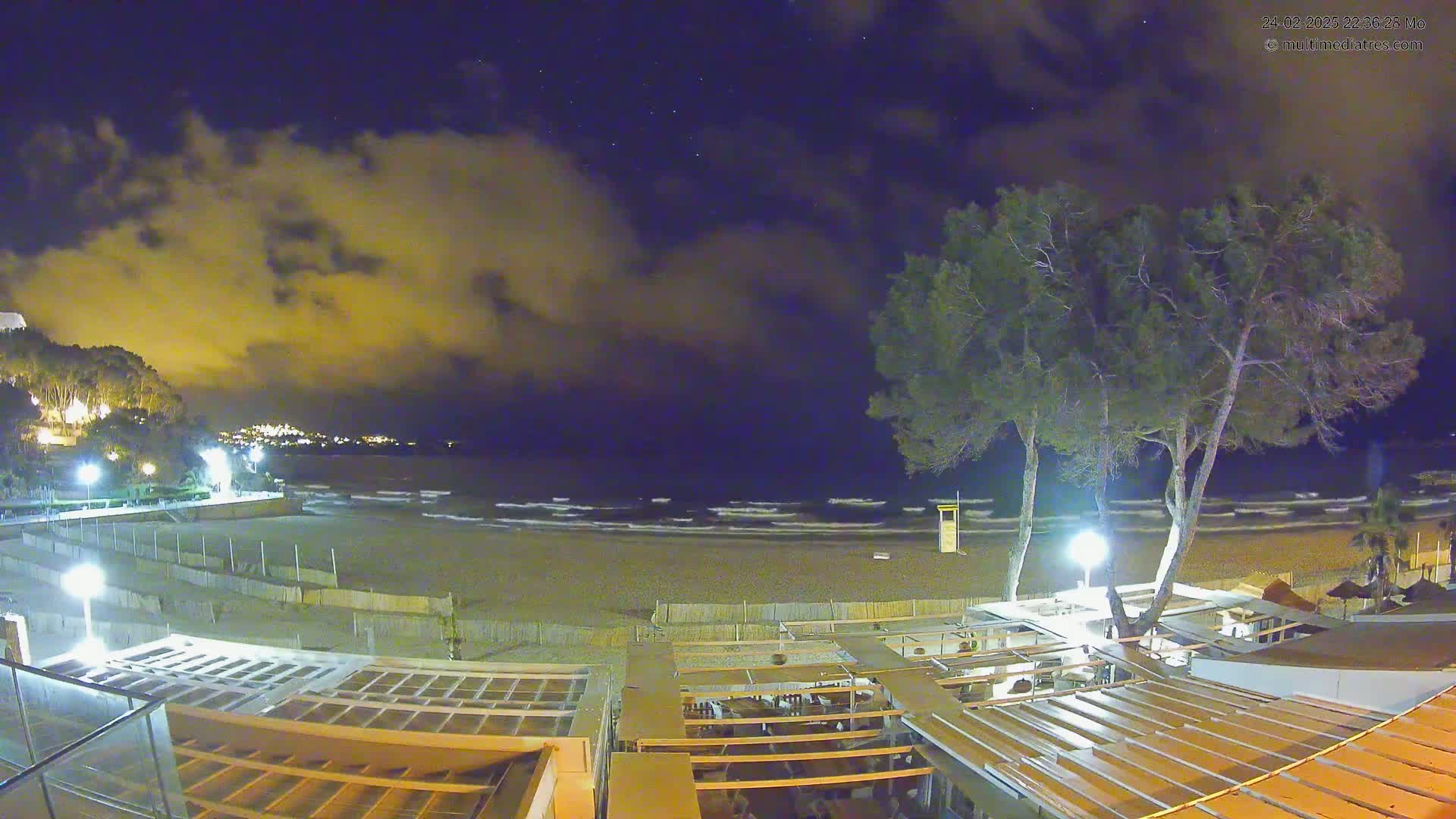 A nighttime view of a beach with gentle waves under a partly cloudy sky, showing a beachfront establishment with outdoor seating areas illuminated by lights.
