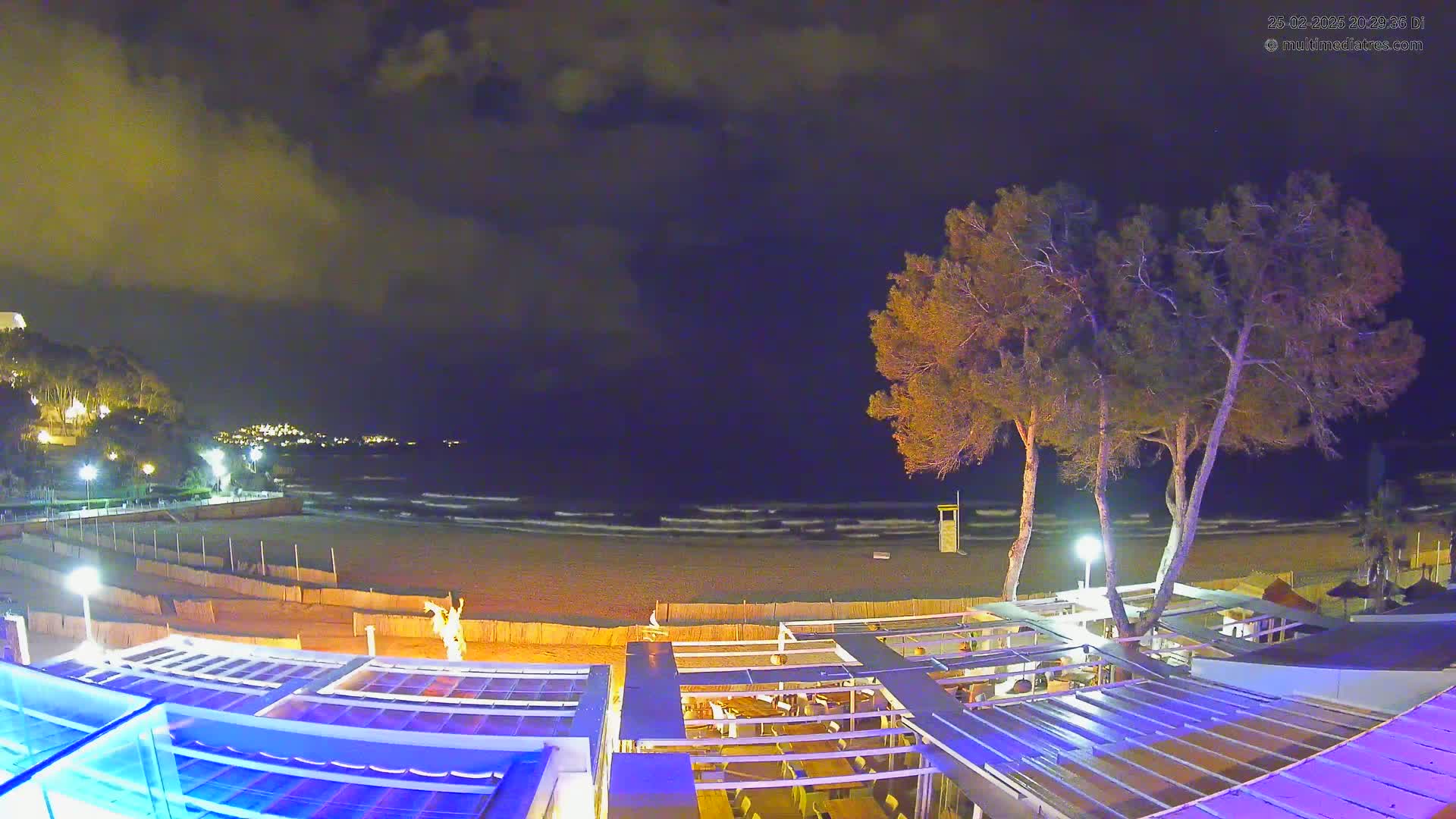 A nighttime view of a beach with gently lapping waves under a partly cloudy sky, overlooking a beachfront restaurant and a lifeguard stand.
