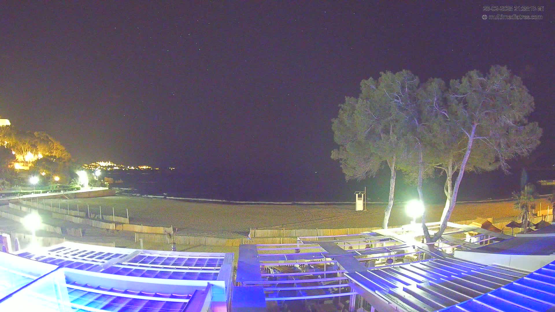 A nighttime view of a beach, with calm water, illuminated trees, and a partially visible building with a blue-lit roof.