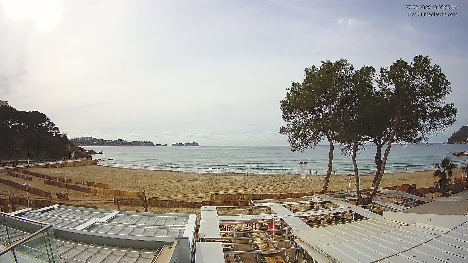 A sandy beach with gentle waves under a cloudy sky is bordered by vegetation, buildings, and a partially enclosed outdoor restaurant area.