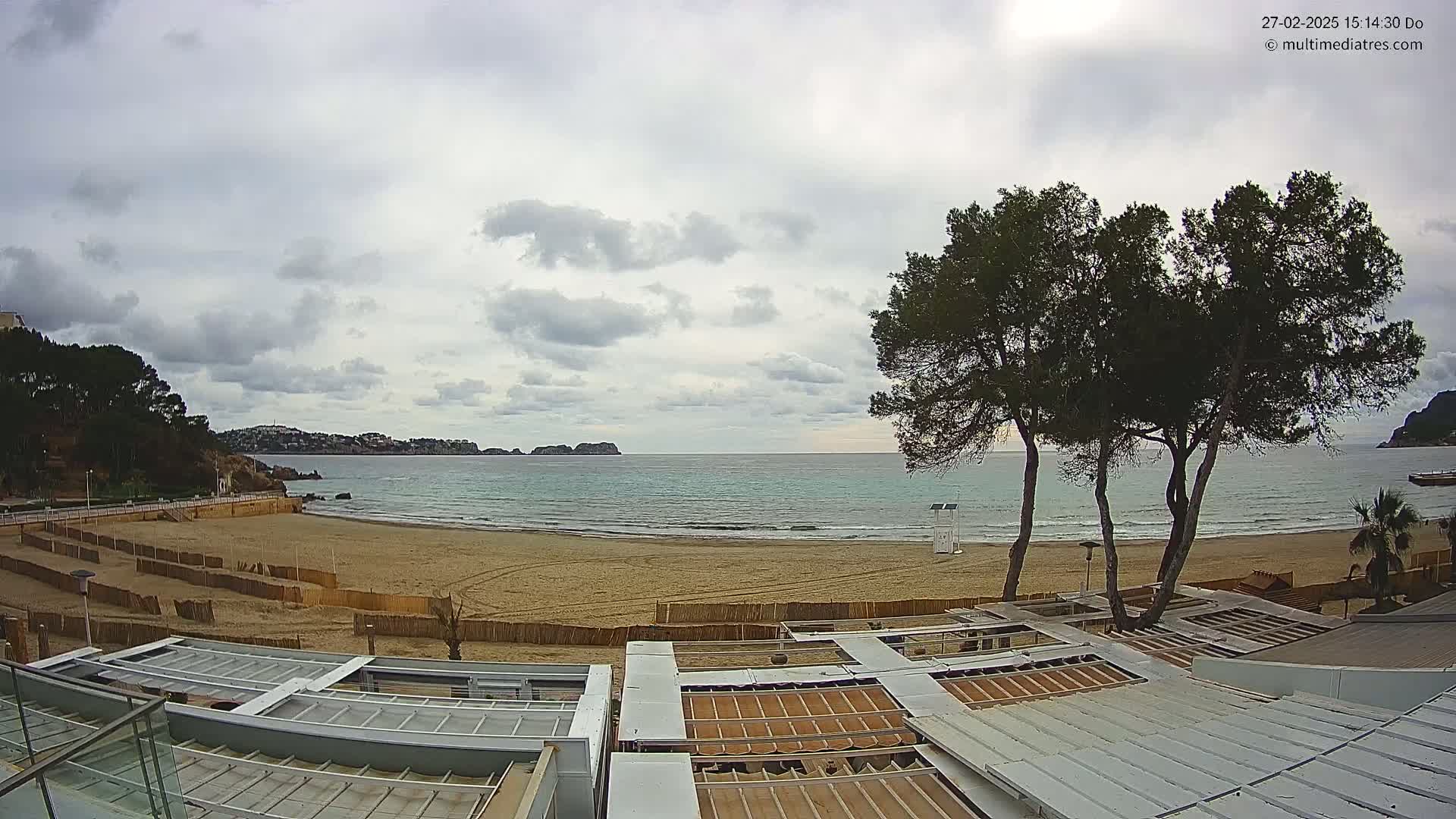 A sandy beach with calm, turquoise water under a cloudy sky is viewed from above a building with retractable roofs, and two tall pine trees stand in the foreground.