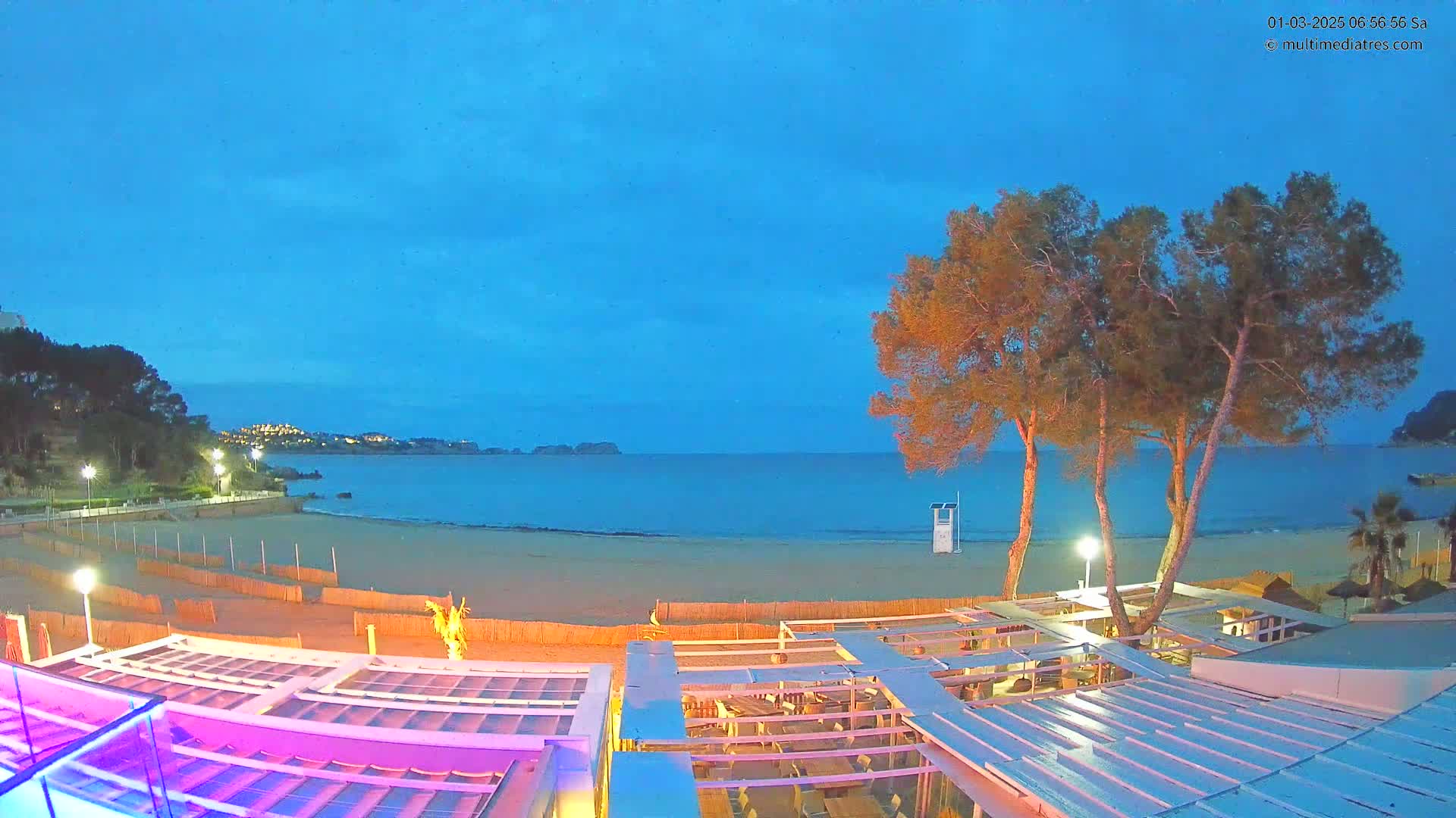 A beach at twilight, under a clear blue sky, is viewed from above a partially lit outdoor restaurant, with several trees visible in the foreground.