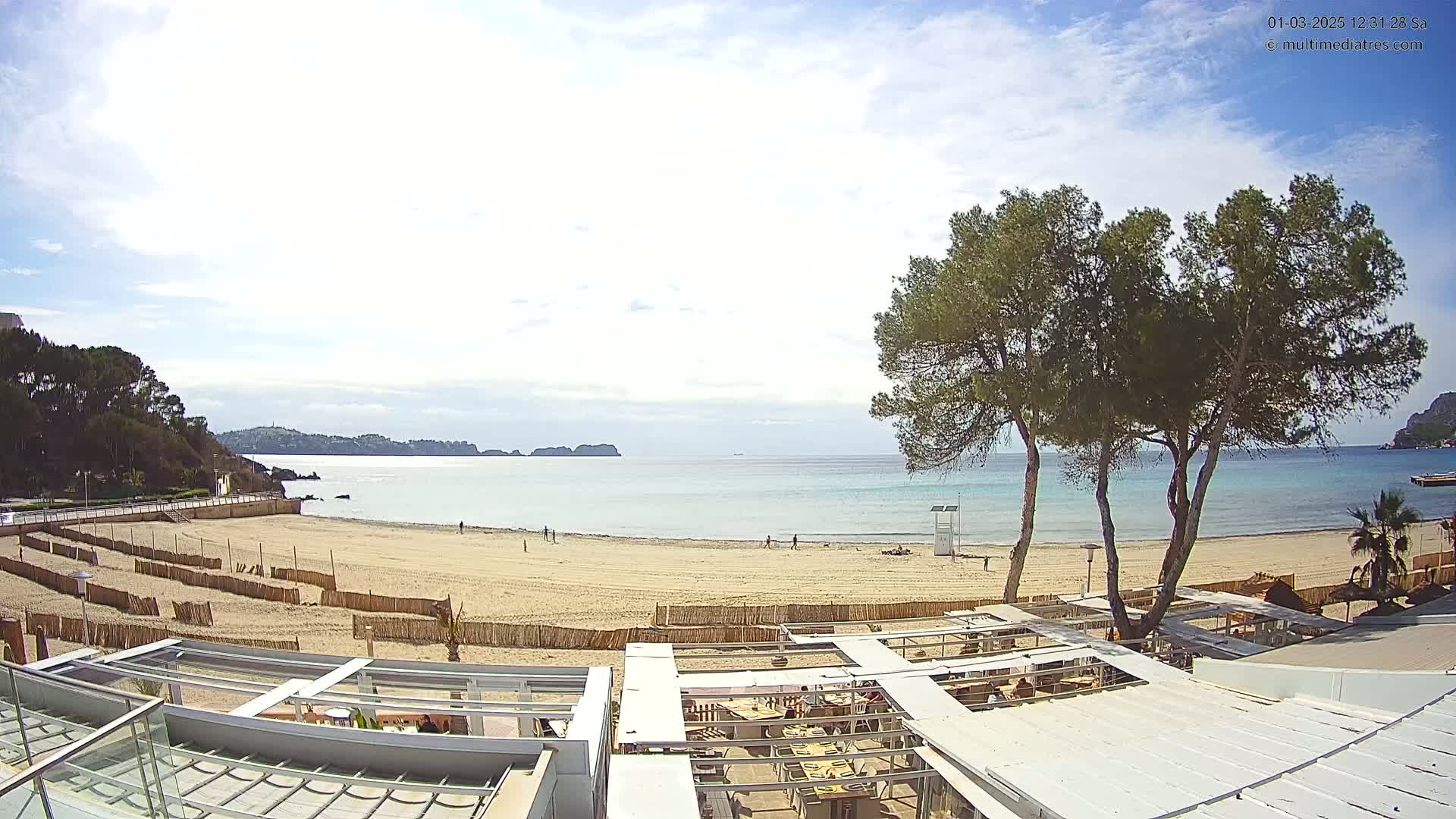 A mostly sunny day at a sandy beach with calm, light-blue water,  a few people,  and several pine trees near a beachfront restaurant.