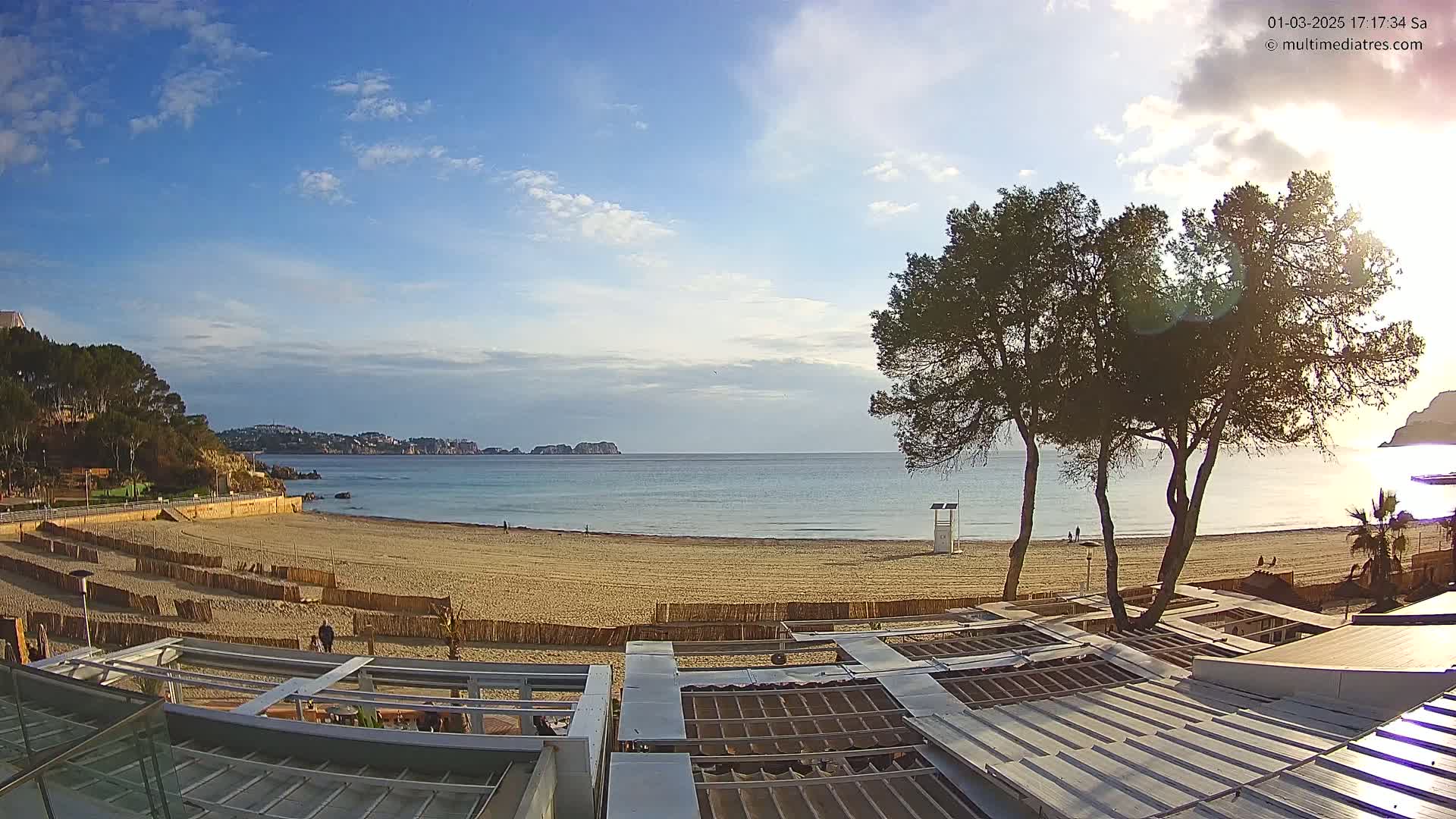 A sandy beach with calm water and a partly cloudy sky is viewed from above a row of beachside structures, with several pine trees in the foreground.