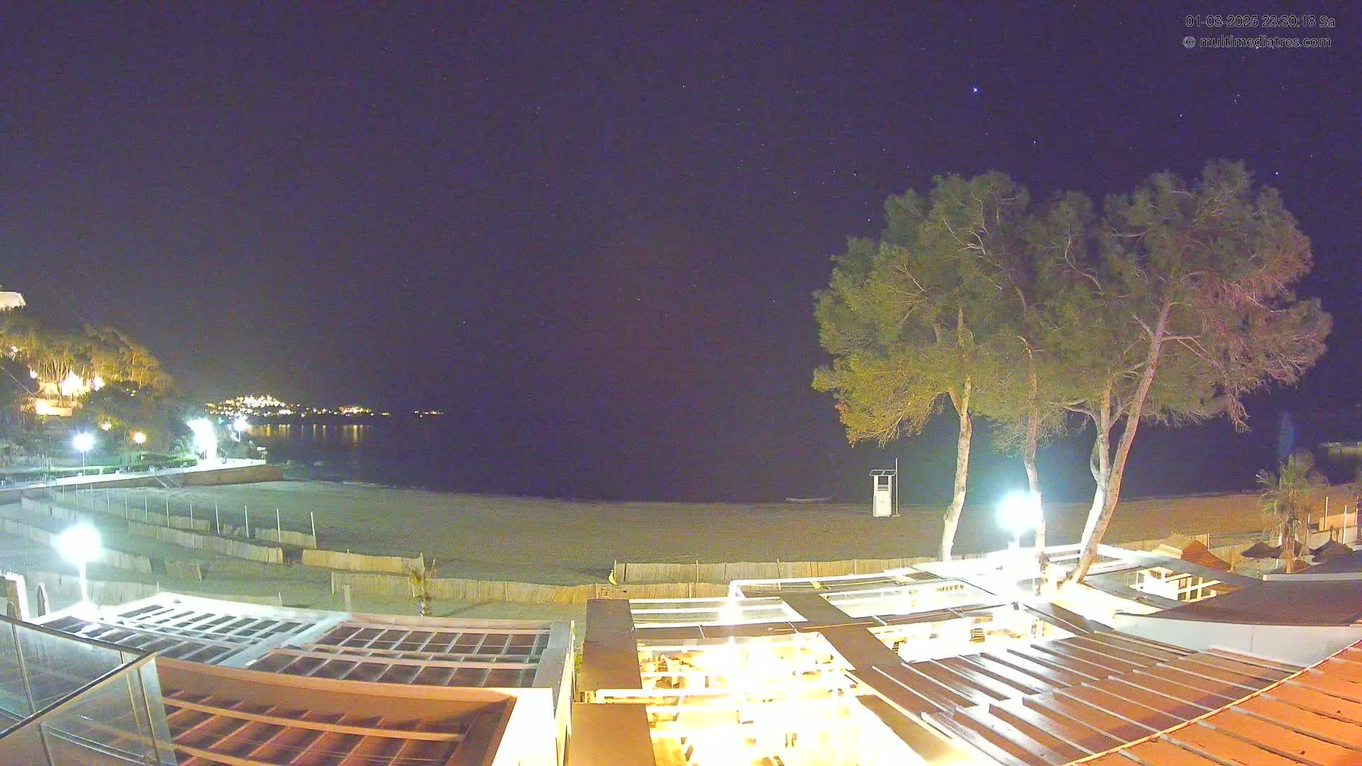 A nighttime view of a beach at the edge of town, under a clear, starry sky, shows a sandy beach, calm water, and several illuminated buildings.