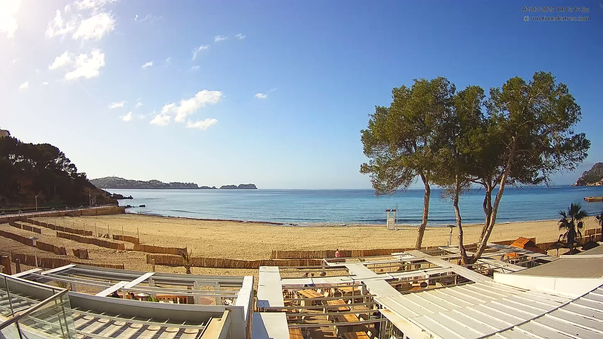 A sandy beach with calm, clear water under a mostly sunny sky is viewed from above a beachfront restaurant with a patio.