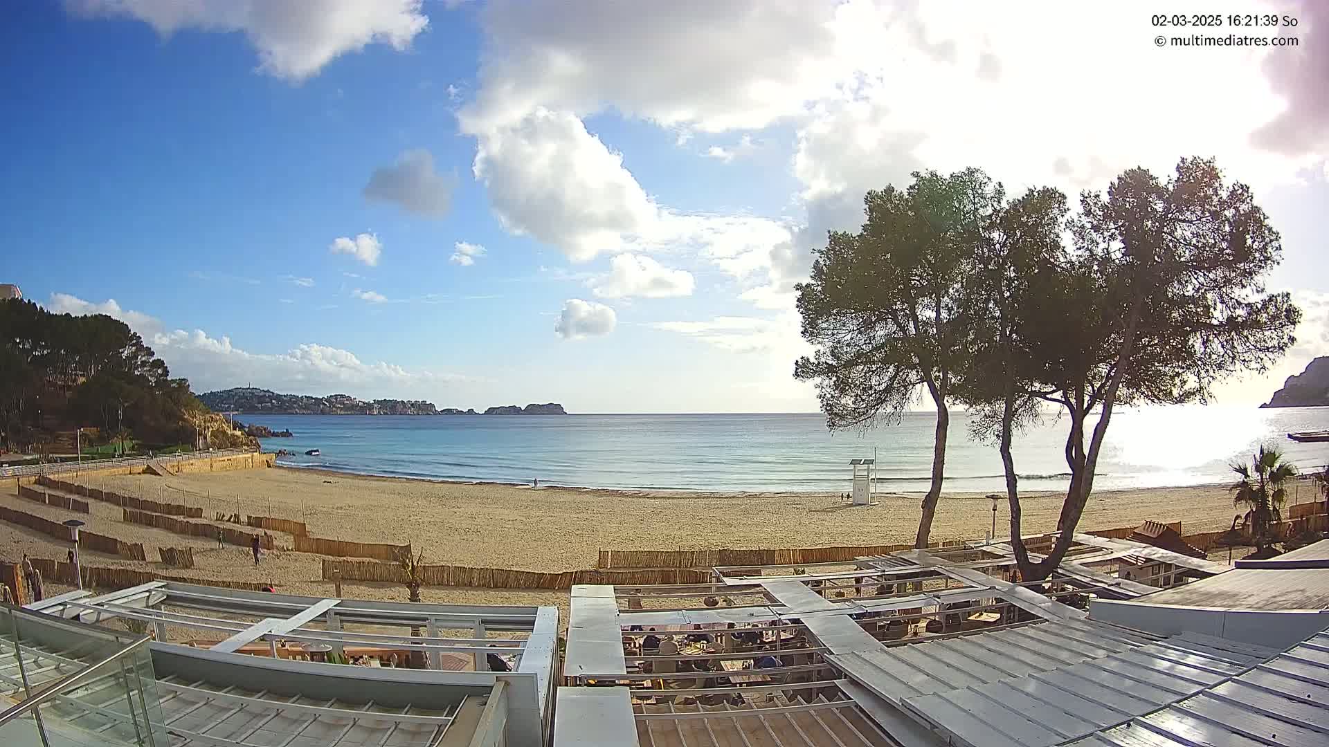 A sandy beach with calm, clear water stretches out beneath a partly cloudy blue sky, viewed from above a beachfront restaurant.