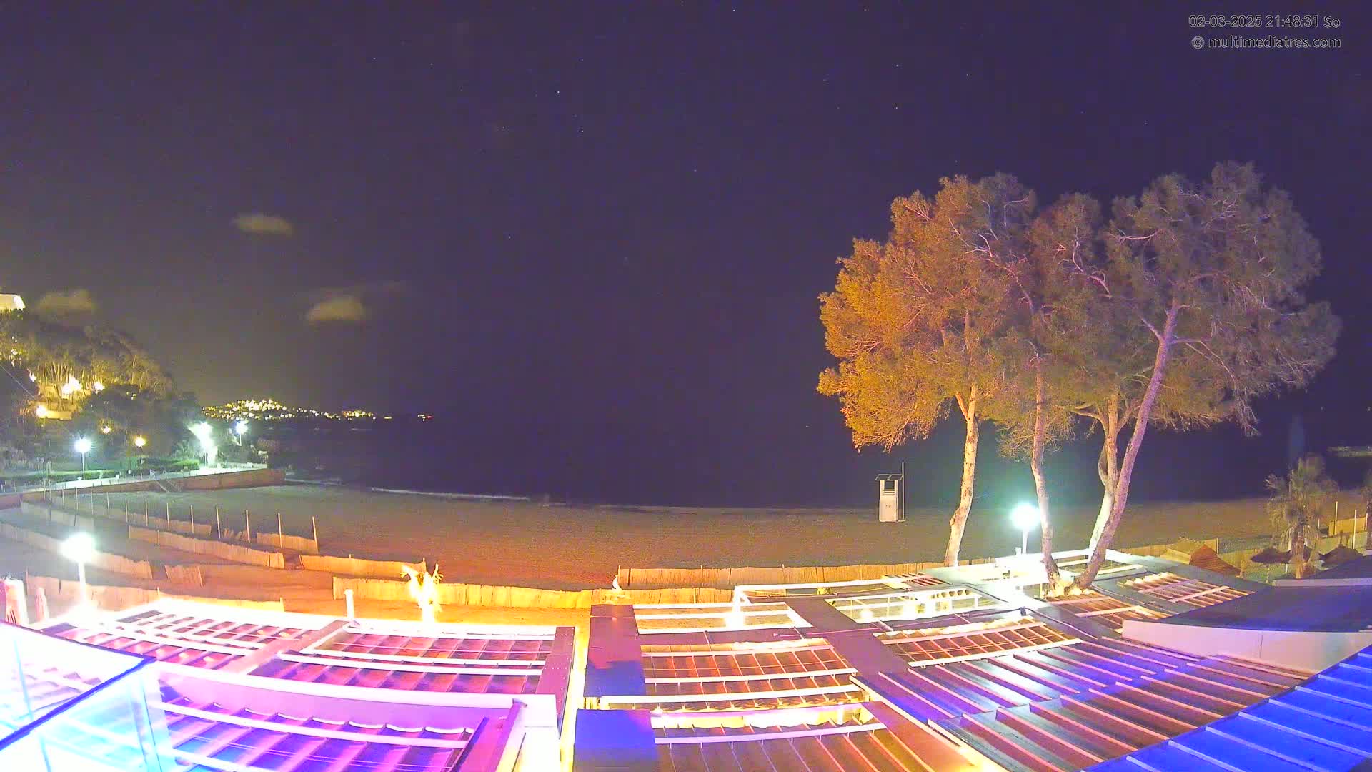 A nighttime view of a beach with a few illuminated buildings and trees under a partly cloudy sky.
