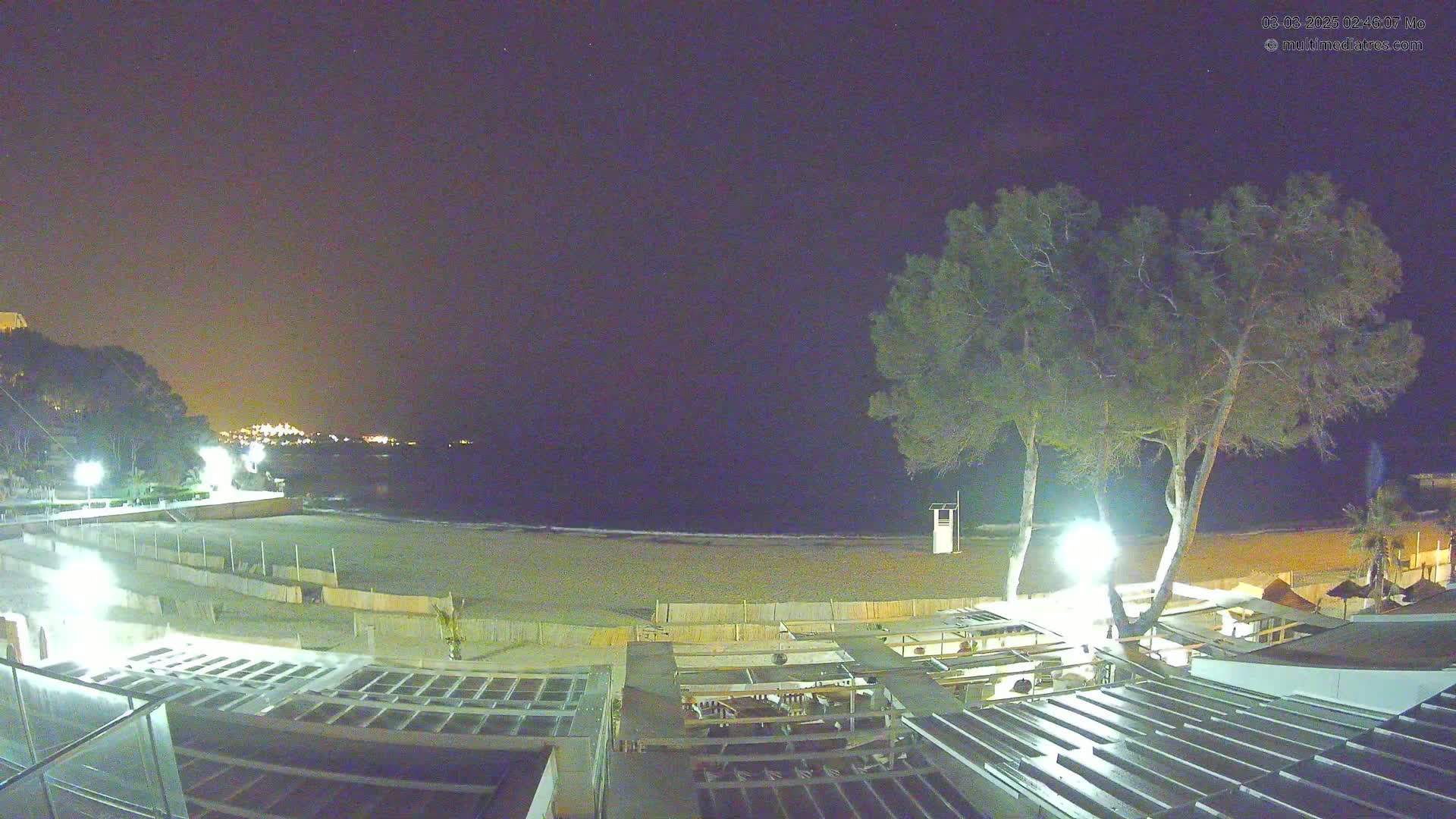 A nighttime view of a beach with a dark sky, illuminated by lights from buildings and trees near the shoreline.