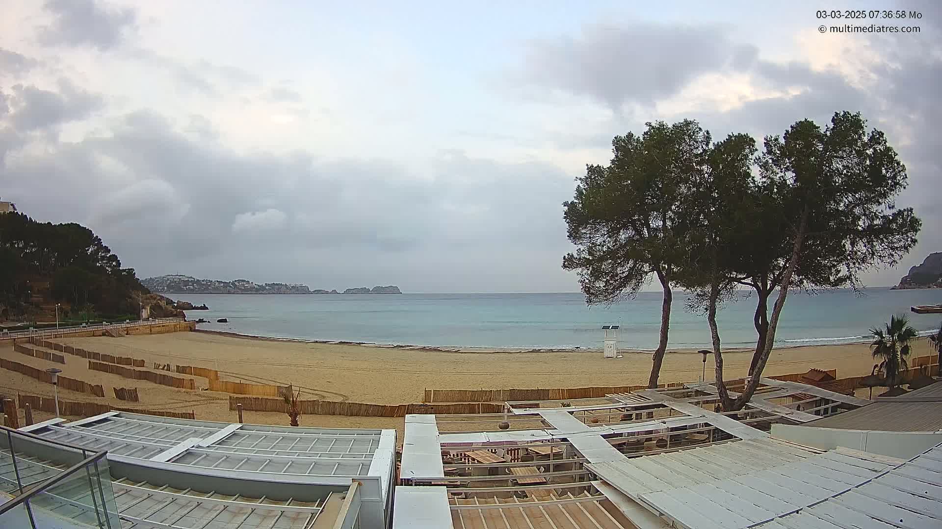 A mostly cloudy day reveals a sandy beach with calm, turquoise water, backed by hills and several tall pine trees near a beachfront structure.