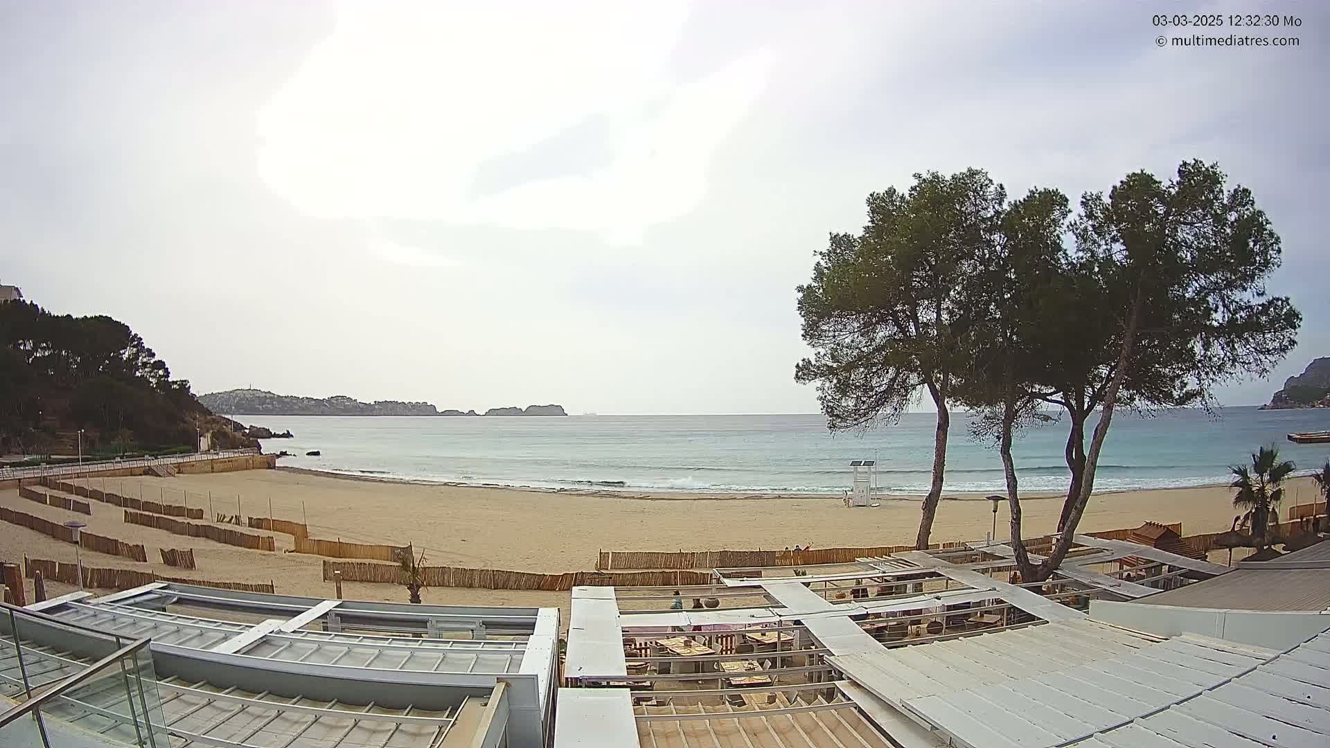 A sandy beach with calm waves and overcast skies is viewed from above a partially covered outdoor restaurant, with green hills and trees in the background.