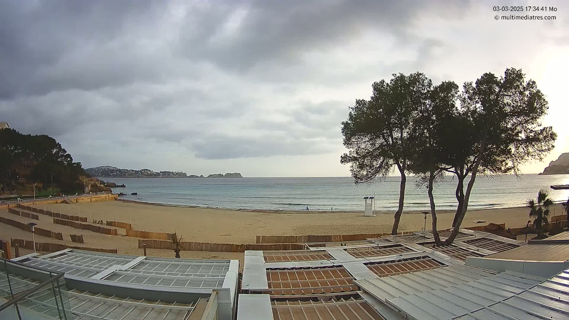 A sandy beach with calm ocean waves under a mostly cloudy sky, viewed from above a row of buildings with retractable roofs.