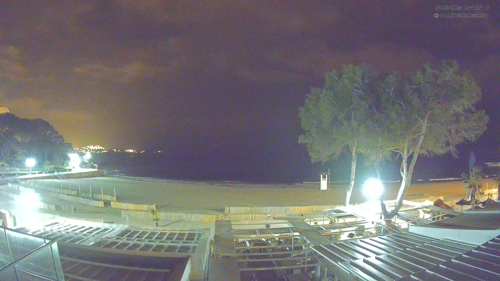 A nighttime view of a beach at low tide with a few lights illuminating a mostly empty sandy expanse, a dark ocean, and several trees near a beachfront building.