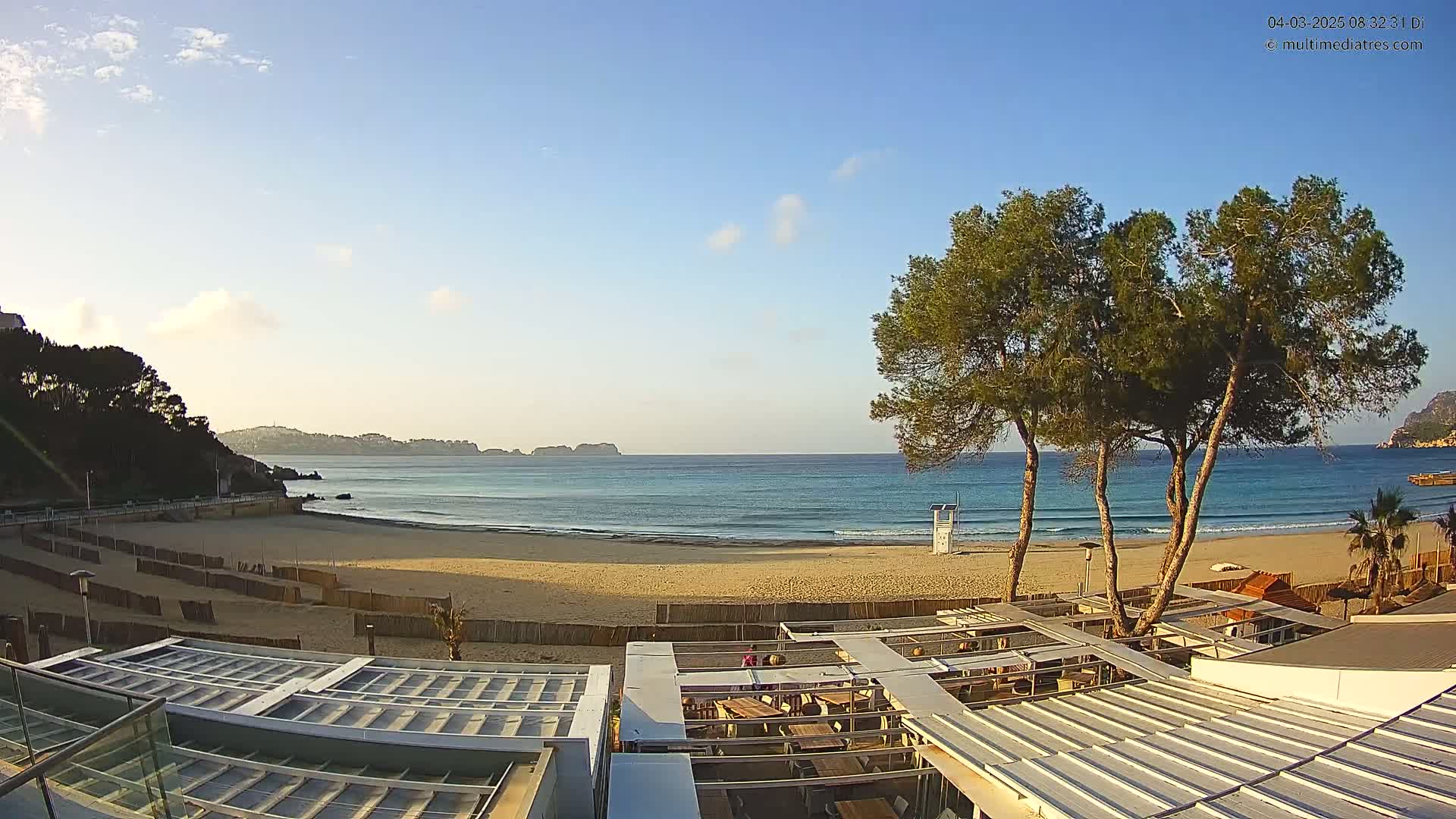 A sandy beach with calm ocean waves is visible under a clear, sunny sky, with several pine trees and a beachside restaurant partially seen in the foreground.