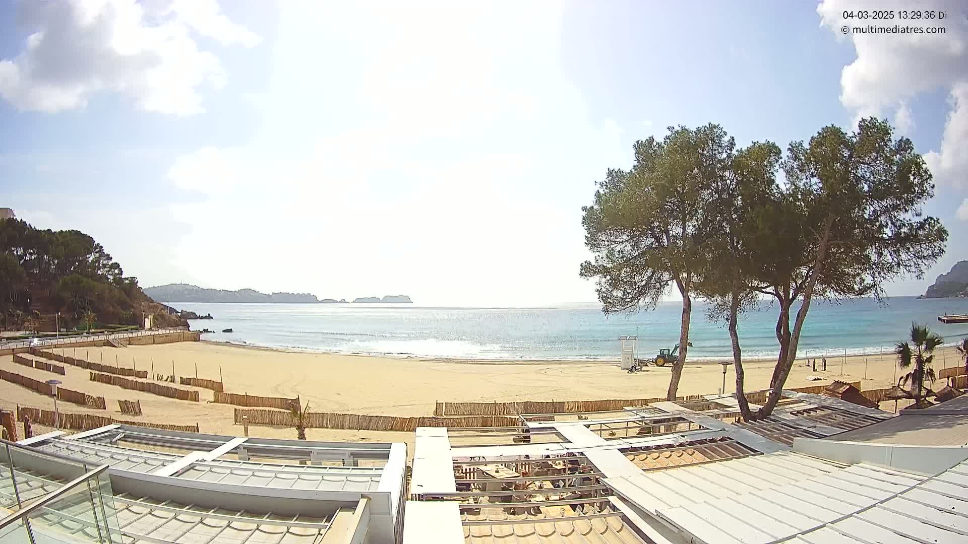 A mostly sunny day at a sandy beach with calm waves, partially enclosed by hills and featuring a few trees and beachside structures under construction.