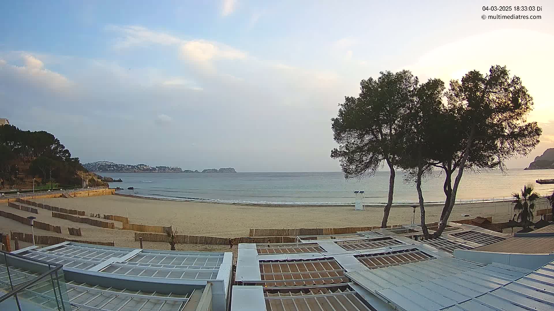 A mostly clear sky overlooks a sandy beach with calm ocean waves, a lifeguard stand, and several trees near a building with a metal roof.