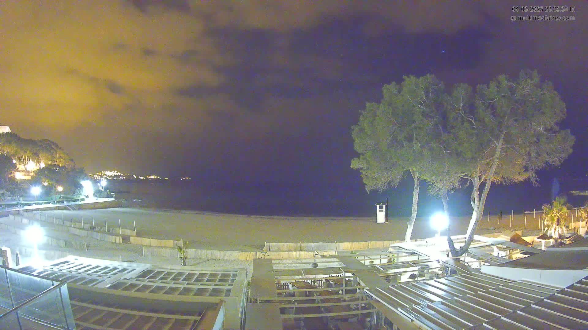 A nighttime view of a beach at low tide, with a partially cloudy sky, showing a beachfront restaurant and two tall trees.