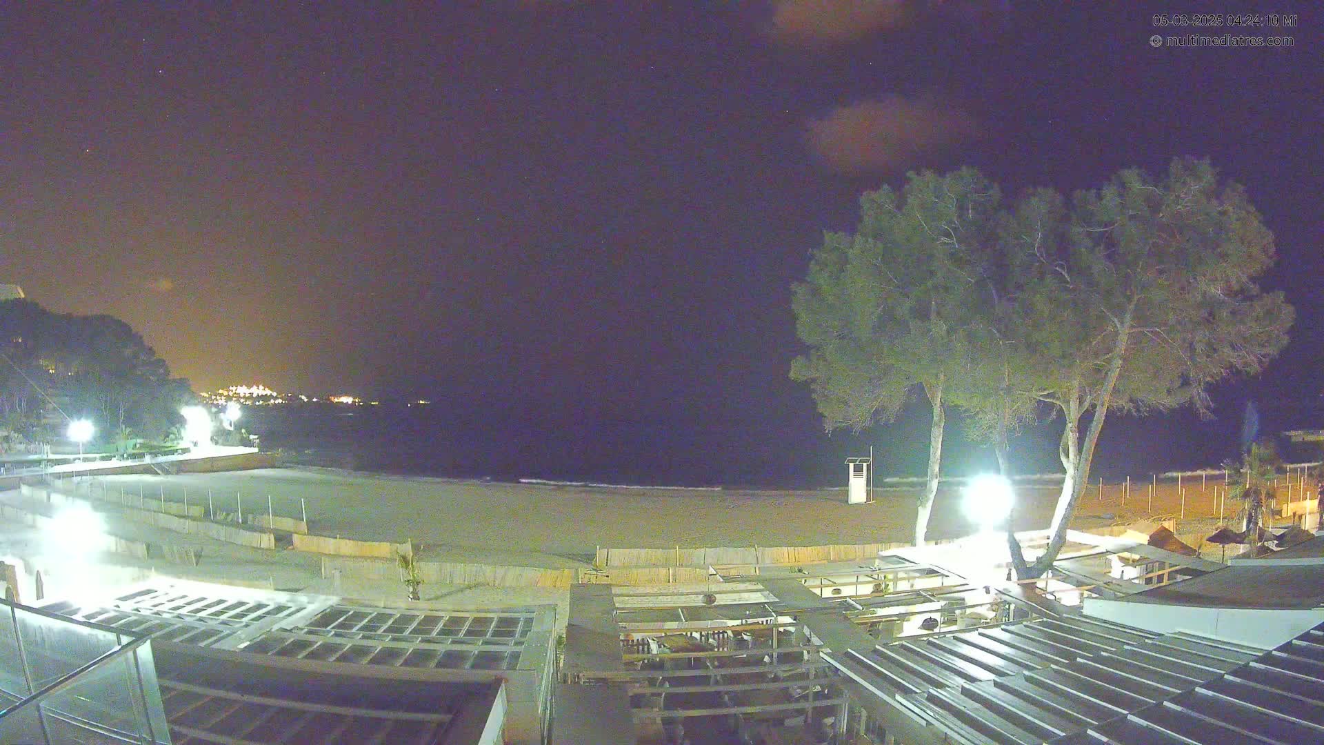 A nighttime beach scene shows a sandy beach, calm ocean, and a beachfront establishment with outdoor seating under the partly cloudy night sky.